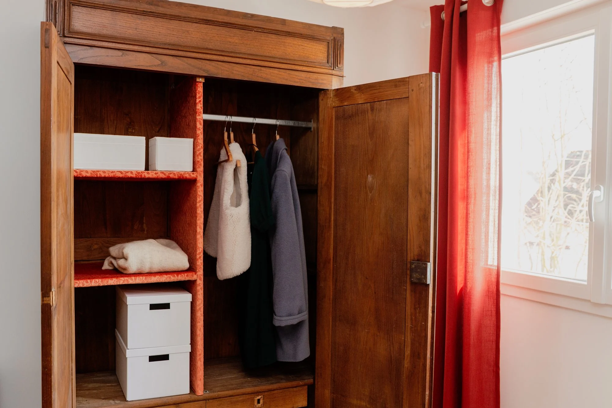 Open wooden wardrobe with hanging coats, folded clothing, storage boxes, and red curtains beside a window