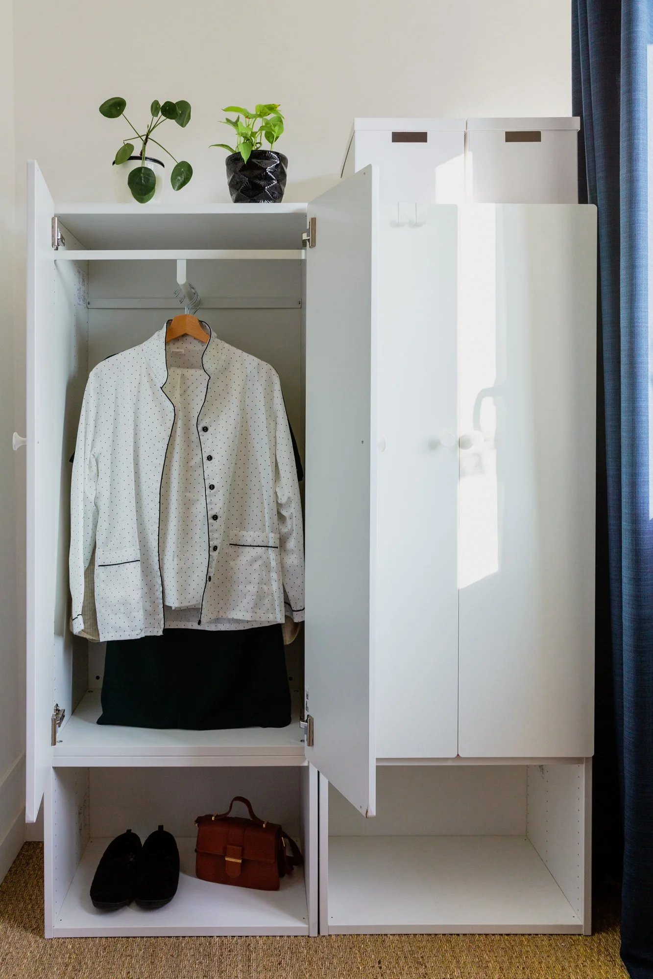 Open white wardrobe with a polka-dot shirt, black pants, shoes, handbag, storage boxes, and potted plants