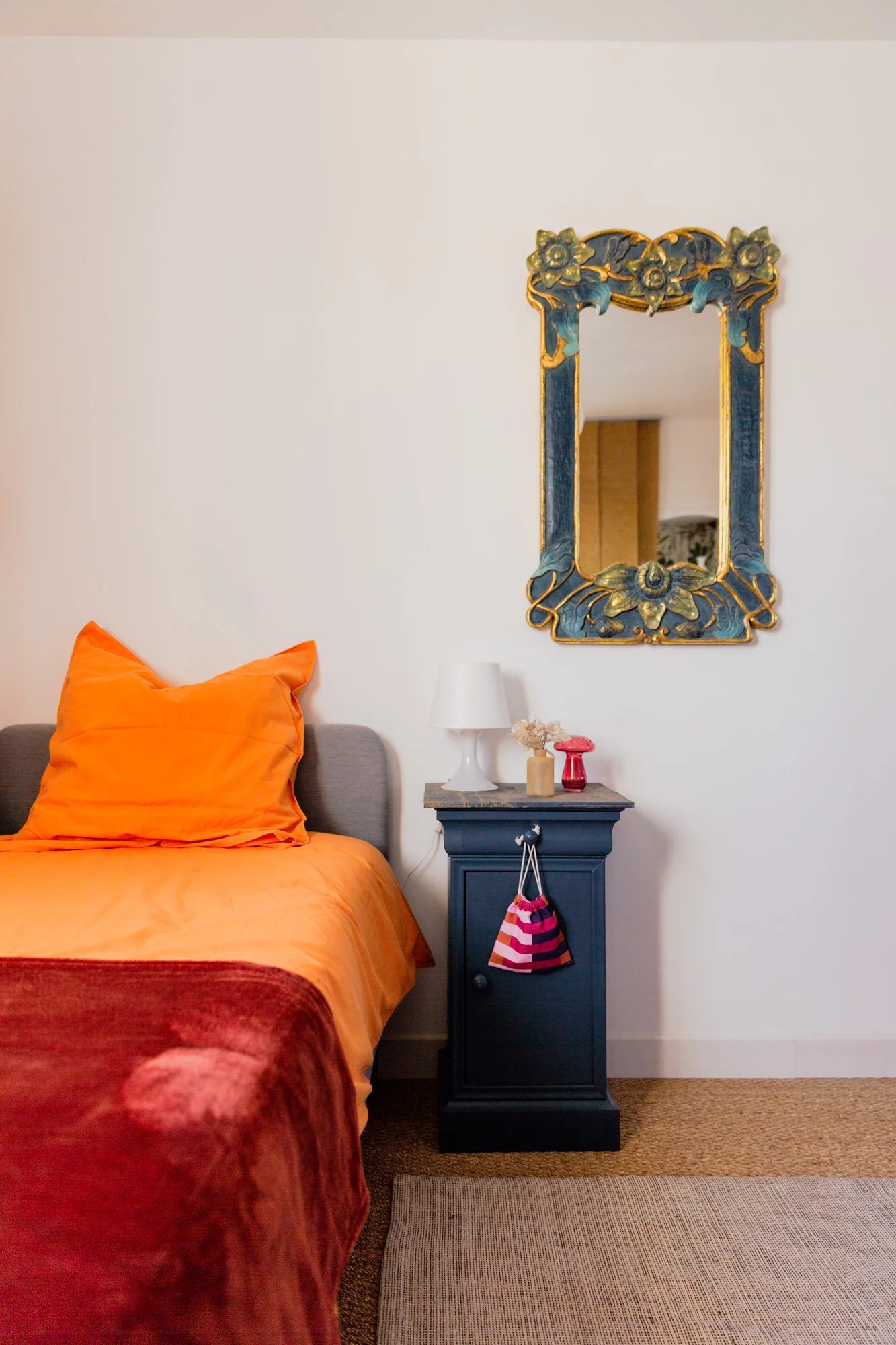 Colorful bedroom with orange bedding, ornate blue-and-gold floral mirror, dark nightstand, white lamp, and decorative vases