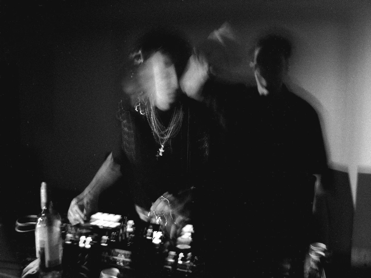 A woman with dark hair and layered necklaces operating DJ equipment, with another person standing nearby, in a dimly lit setting.