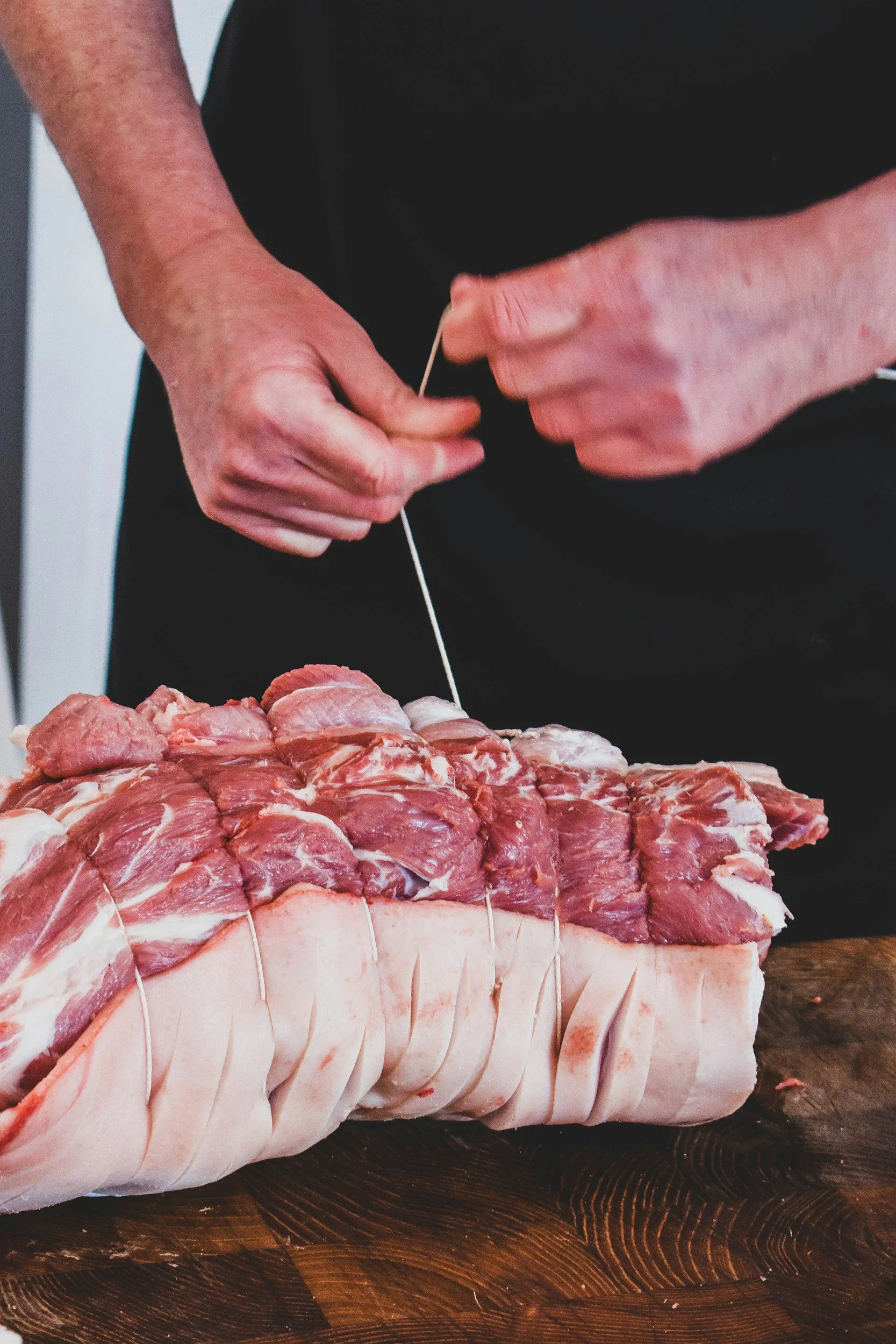 A person preparing a large cut of meat, tying it with string on a wooden cutting board.