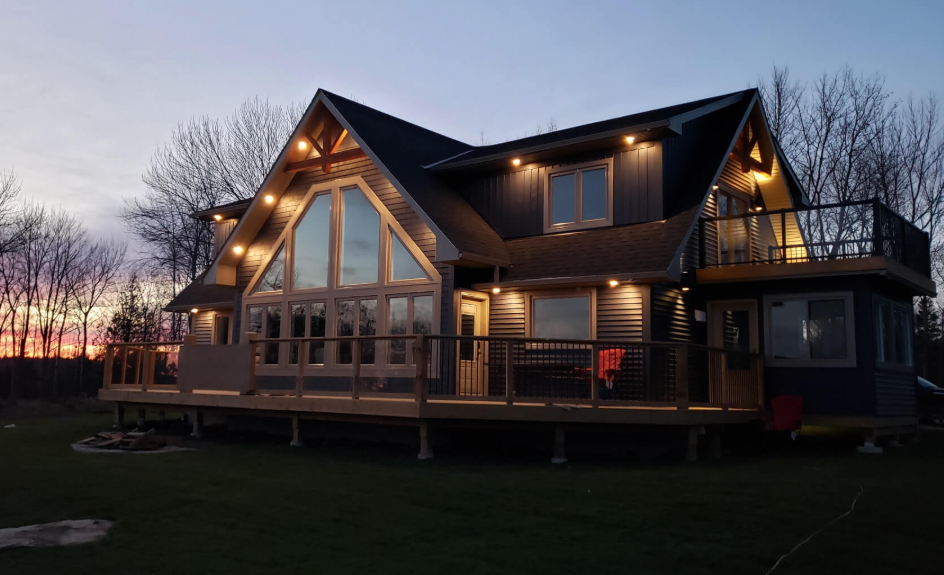 A modern two-story house with large front windows, a deck, and exterior lighting at dusk, surrounded by bare trees.