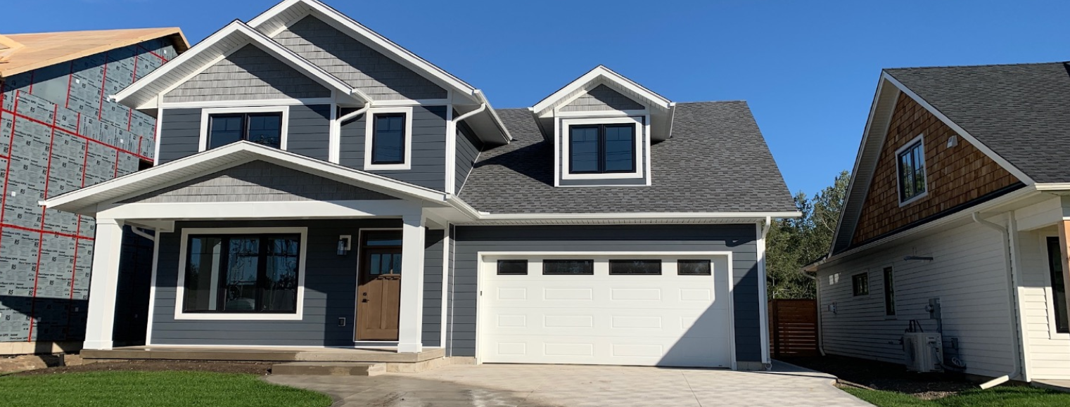 New gray house with a two-car garage, white trim, front porch, brown front door, and dormer windows, beside a white house under construction, on a clear day.