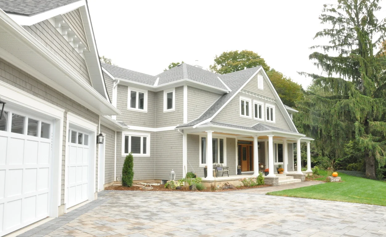 Front view of a large house with a gray exterior, white trim, and a covered porch, surrounded by a landscaped yard with a stone driveway and large trees in the background.