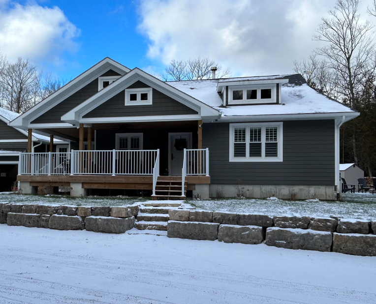 A modern house with dark gray siding, white trim, a large front porch with white railing, and a snow-covered roof, surrounded by snow on the lawn and steps.