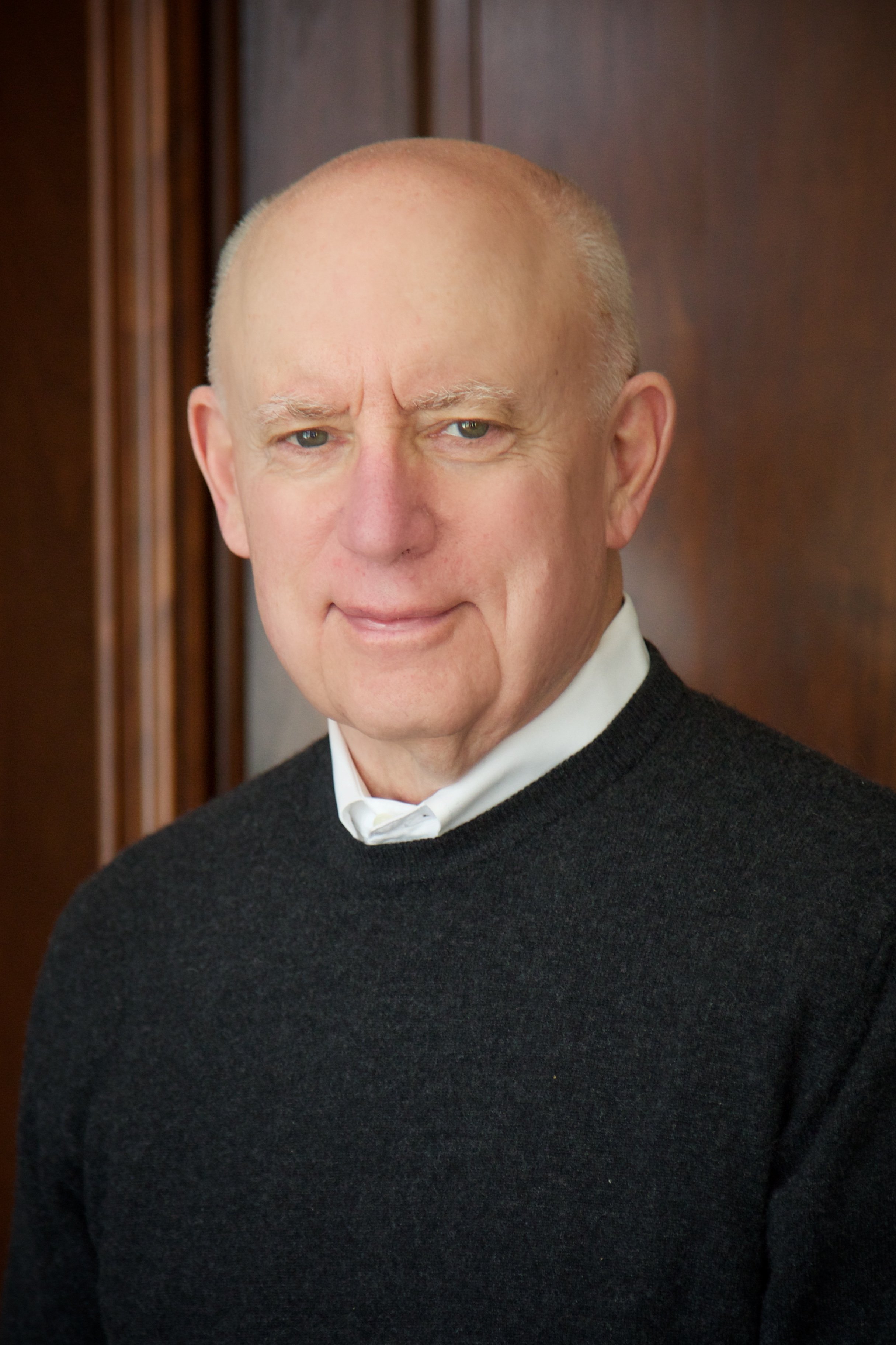 Close-up of a bald man with light skin, wearing a black sweater and white shirt, standing in front of a wooden background.