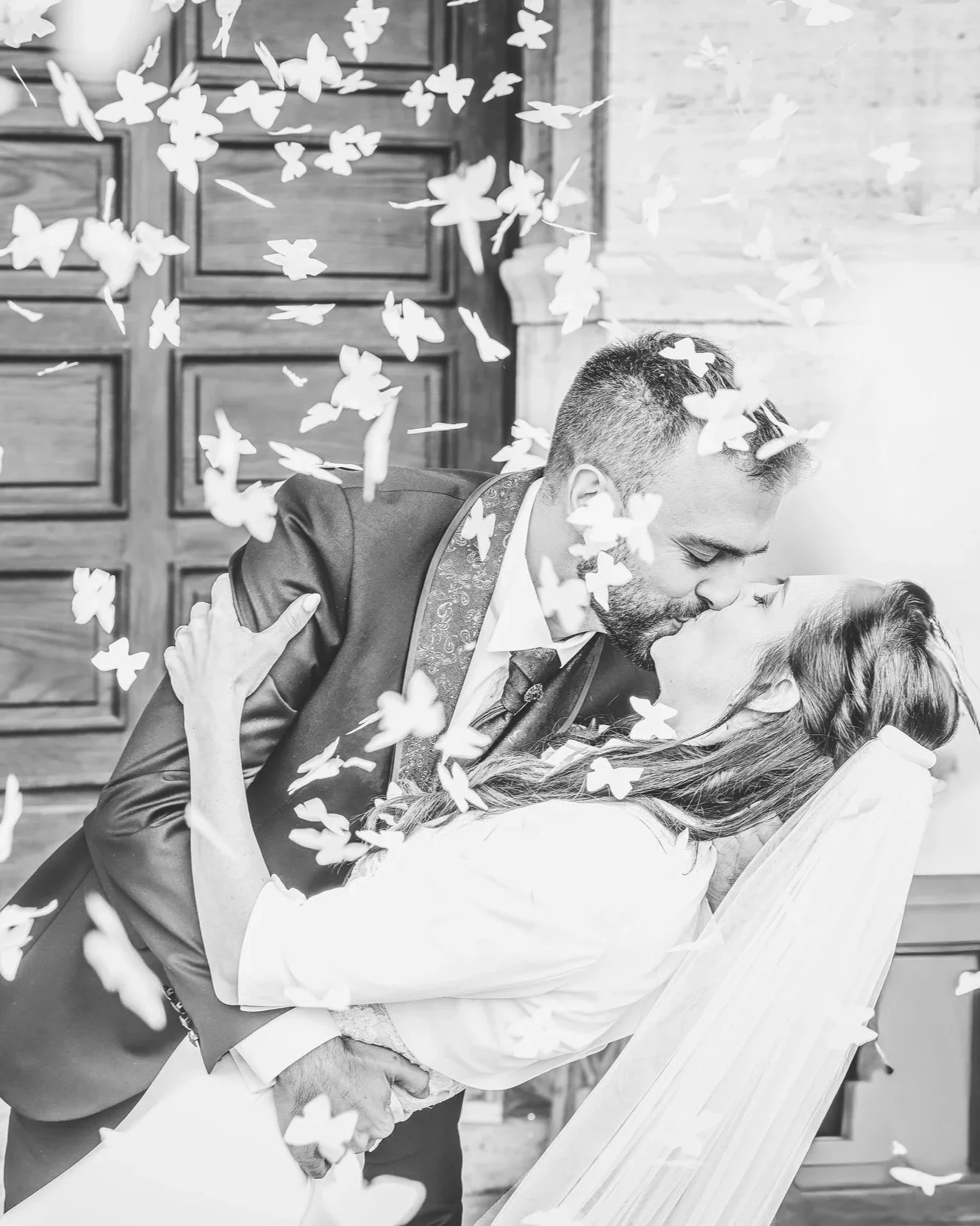 Un uomo e una donna si stanno baciando appassionatamente con petali di fiori che cadono intorno a loro, in un ambiente interno con pareti di legno.