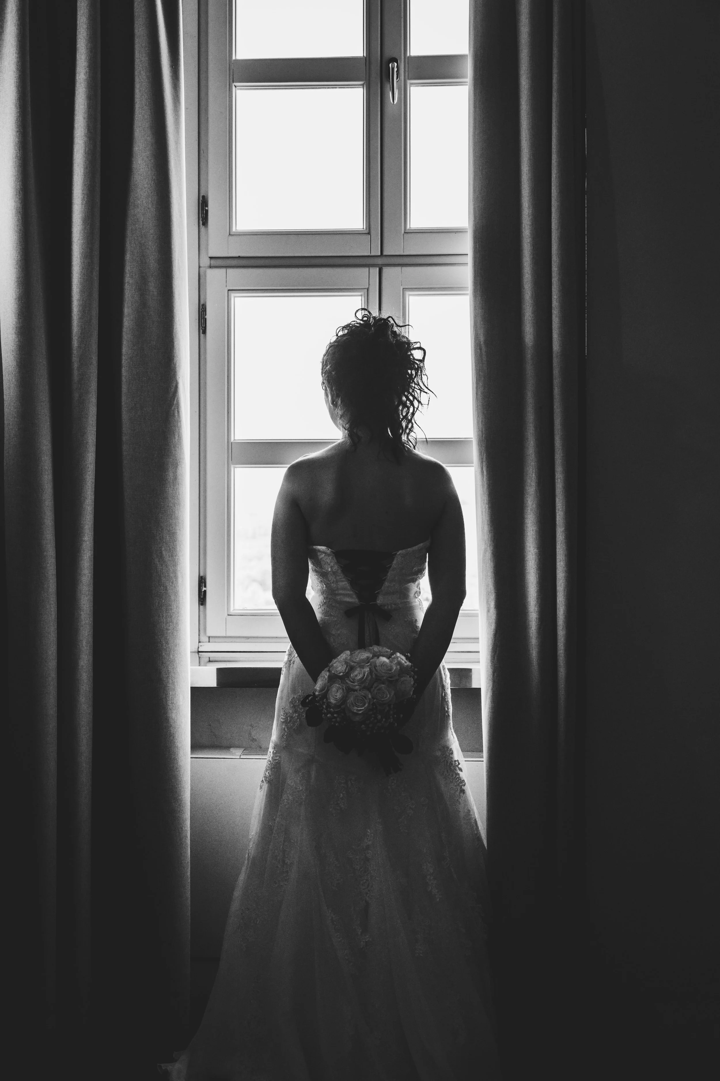 Una donna in abito da sposa si trova davanti a una finestra, con le spalle rivolte verso l'obiettivo, che tiene un mazzo di fiori.