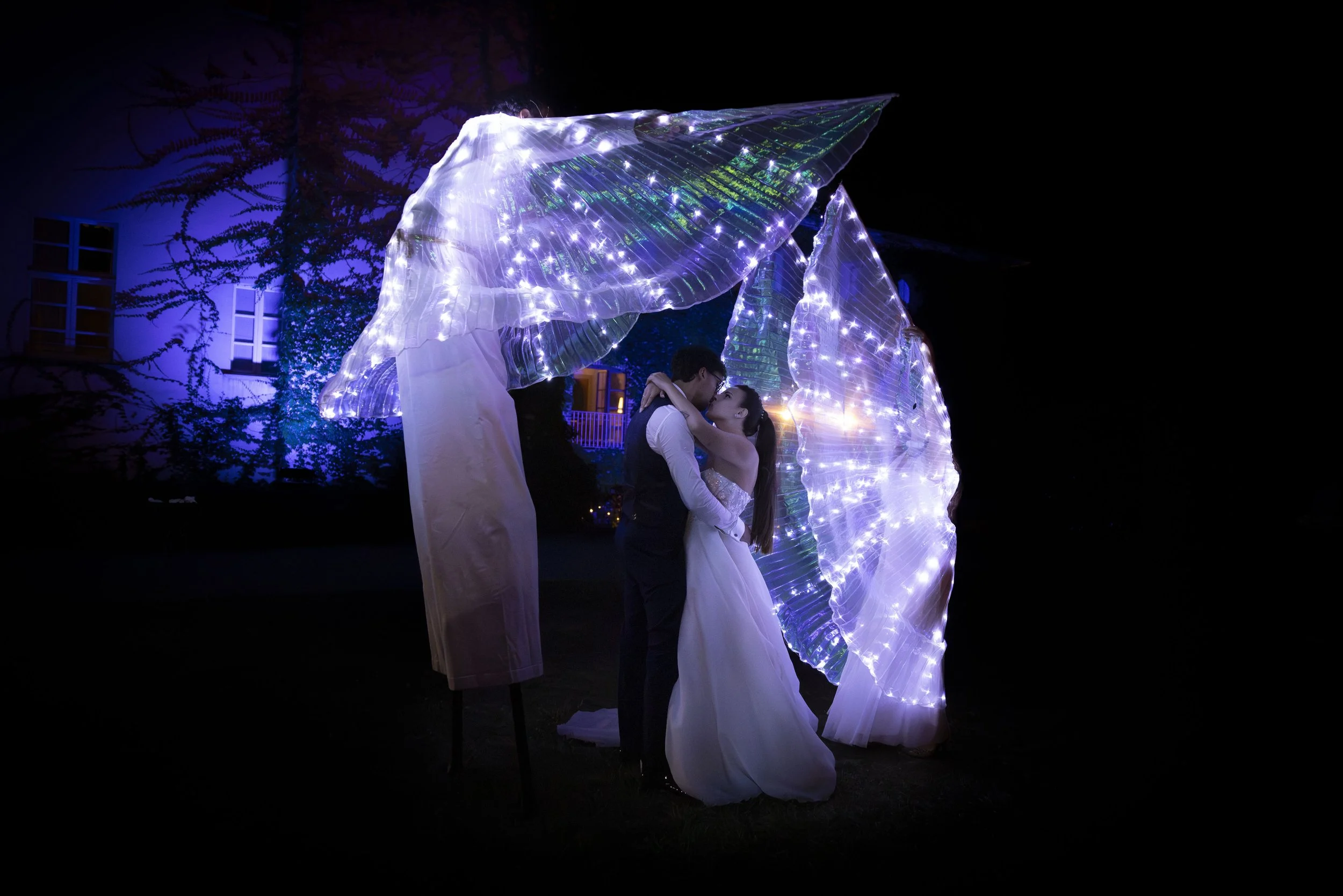 Coppia di sposi che si bacia sotto un'installazione luminosa in seta trasparente illuminata, in un cortile di sera.