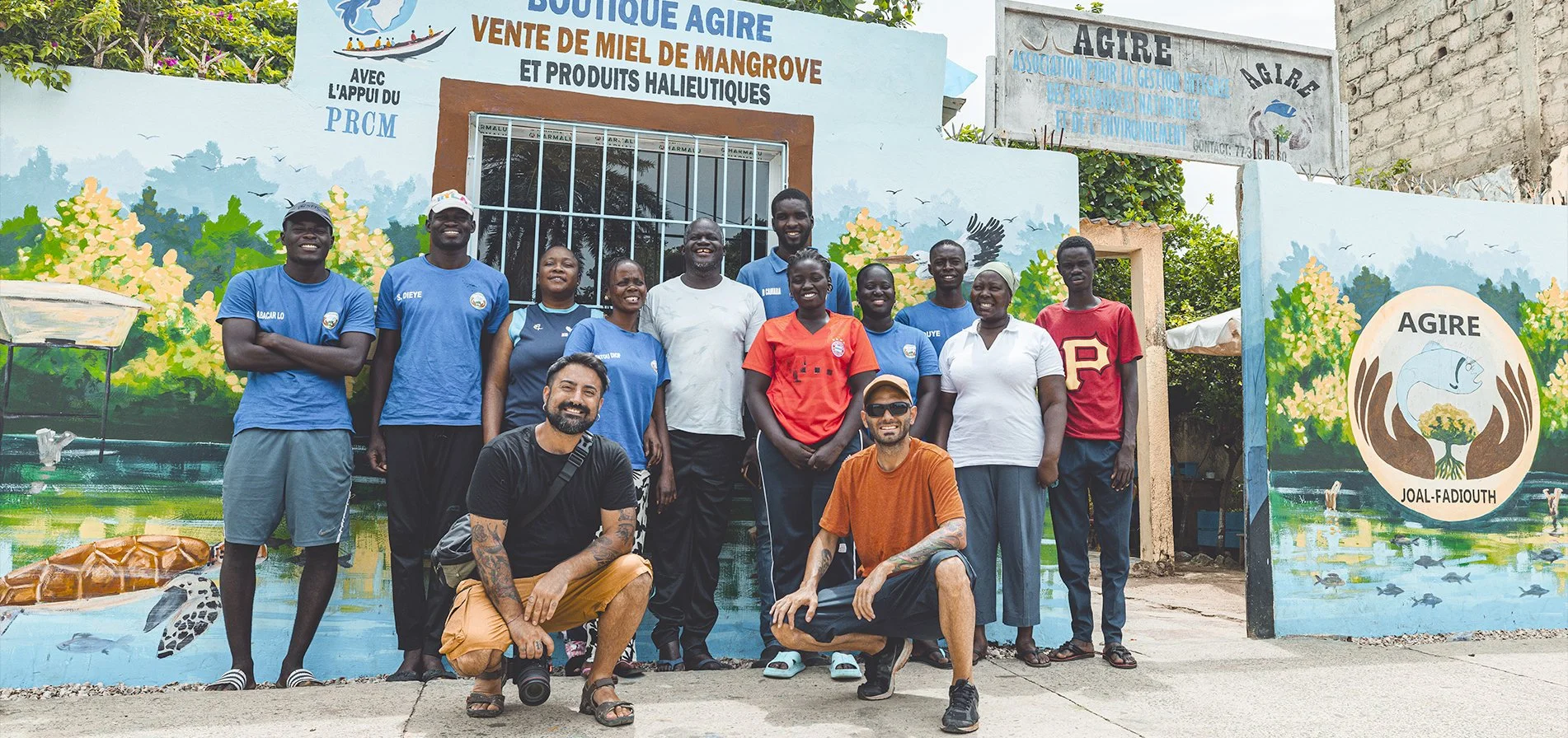 Gruppo di persone davanti a un murale colorato che promuove una vendita di miele e prodotti dell'apicoltura, con scritto 'Vente de miel de mangrove et produits halieutiques' e il logo di 'Agire Joa-Fadiouth'.