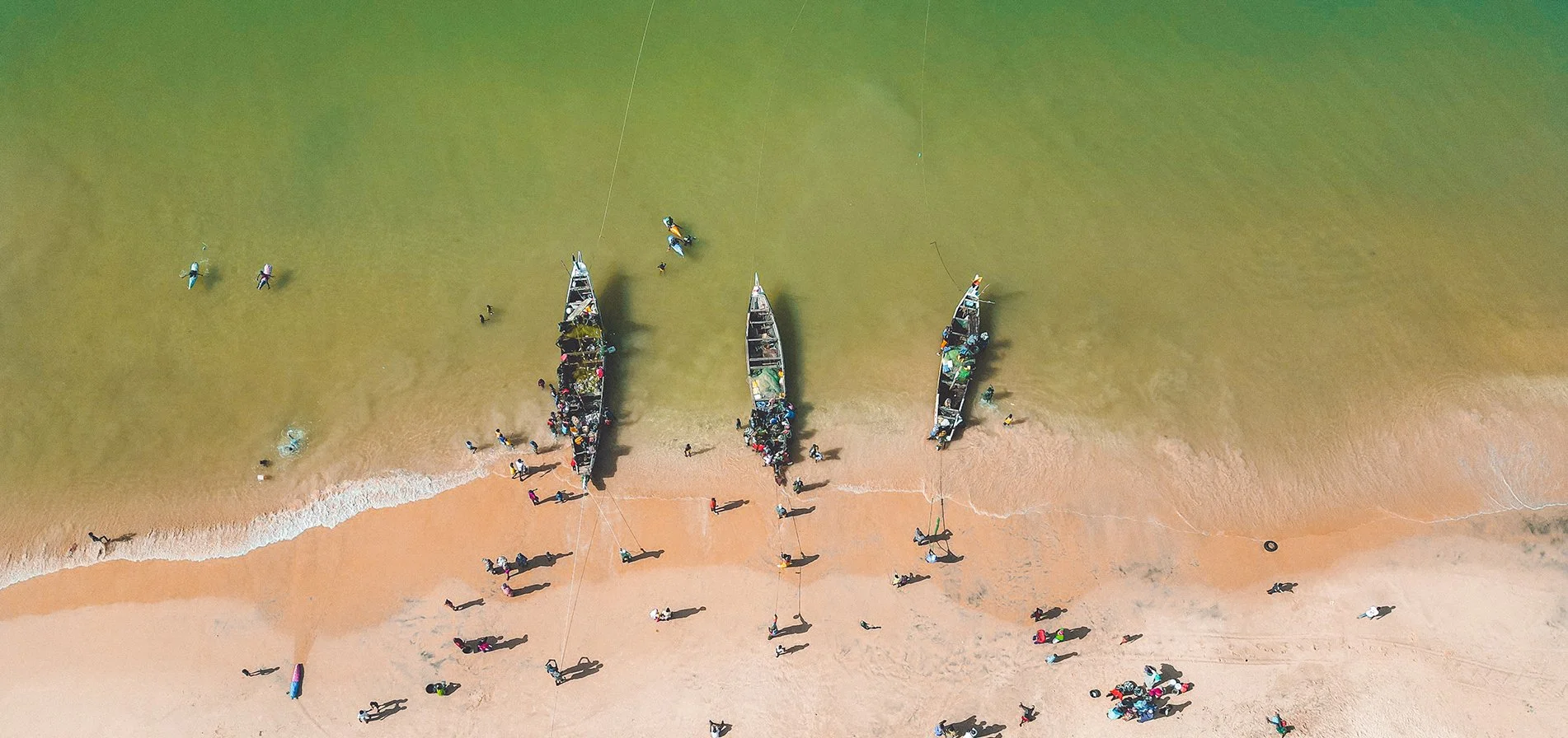 Immagine aerea di una spiaggia con quattro barche alzate sulla sabbia e molte persone che camminano o nuotano nell'acqua