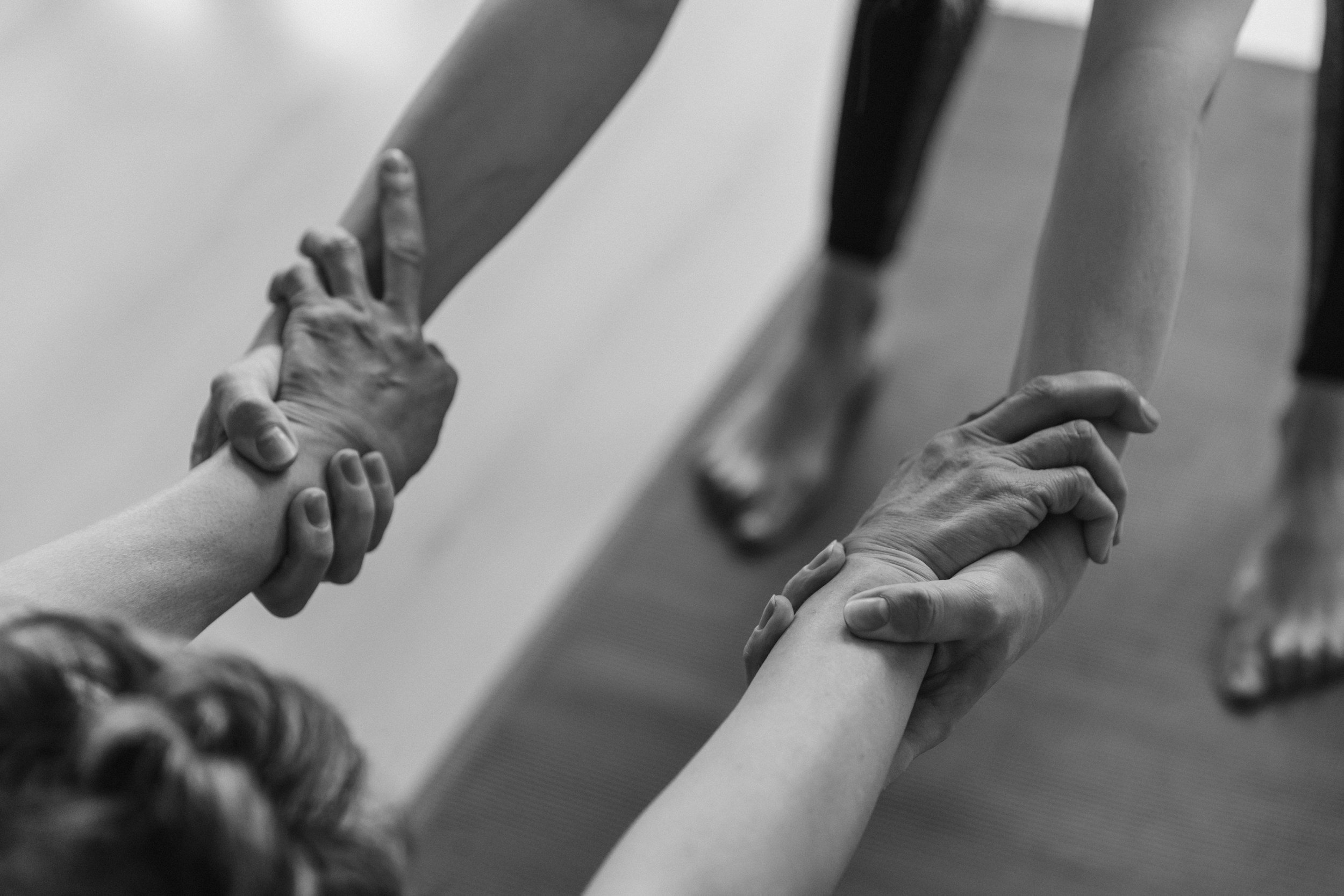 Two people holding hands during a yoga or exercise session, one person providing support with a gentle grip on the other's wrist and hand, on a yoga mat.