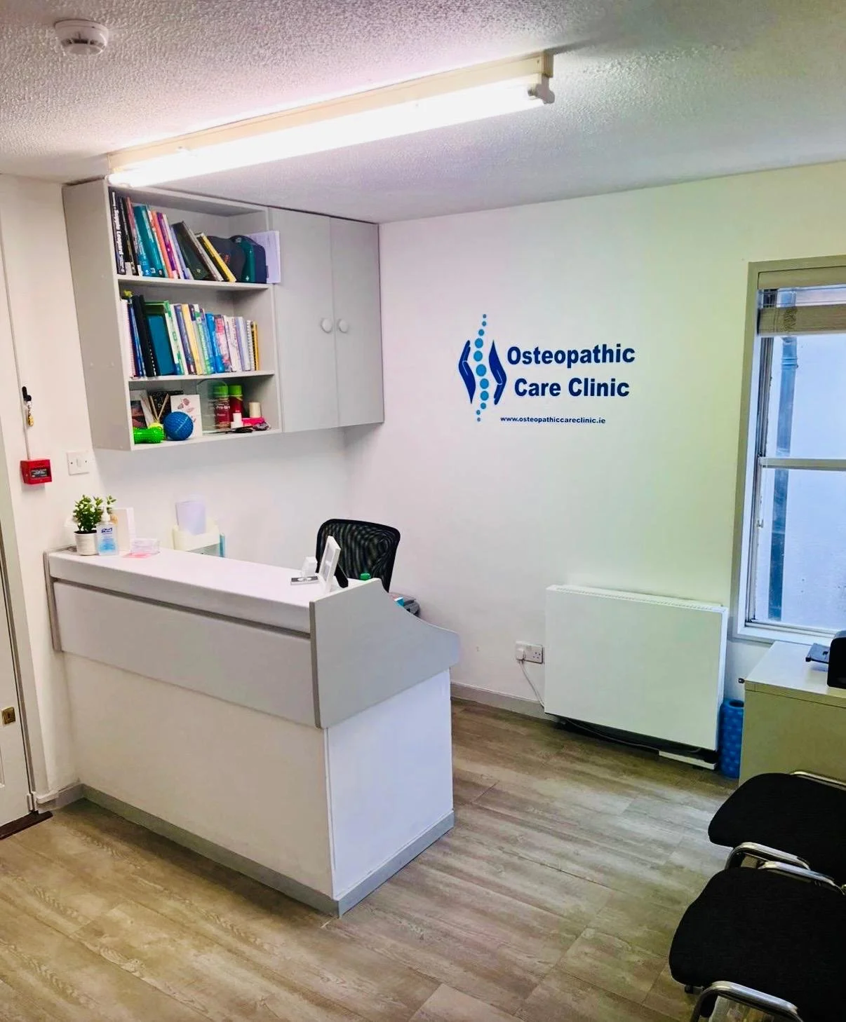 Interior of Osteopathic Care Clinic, featuring a white reception desk, a black office chair, wall-mounted bookshelf with books and objects, clinic logo on the wall, window, and a couple of chairs for waiting.