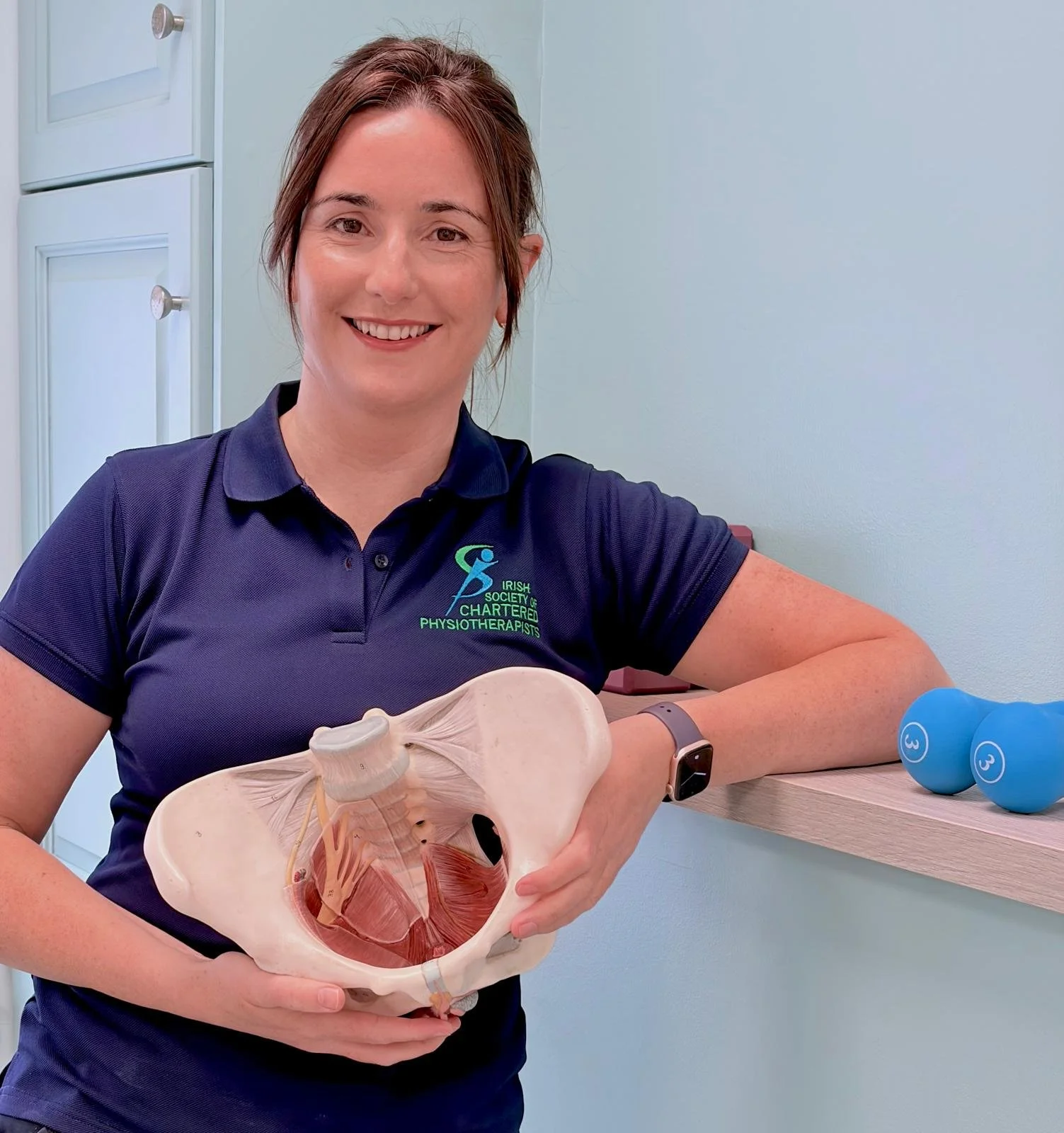 A woman in a navy polo shirt holding a pelvis model and smiling, with two blue dumbbells on the wooden shelf nearby.