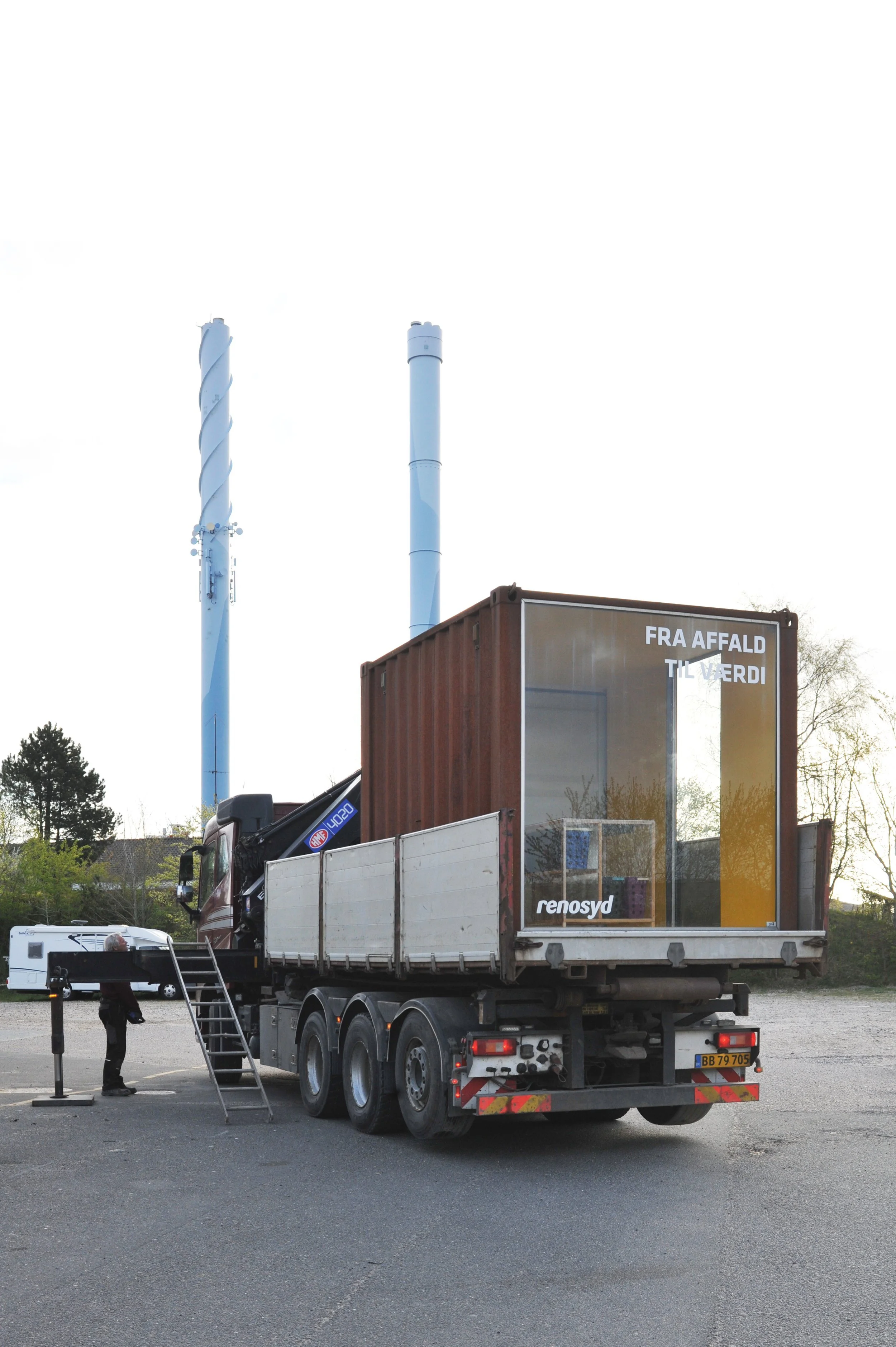 En lastbil med en container, der har skiltet 'Fra affald til værdi' og 'renosyd', står i en parkeringsplads med to store skorstene i baggrunden.