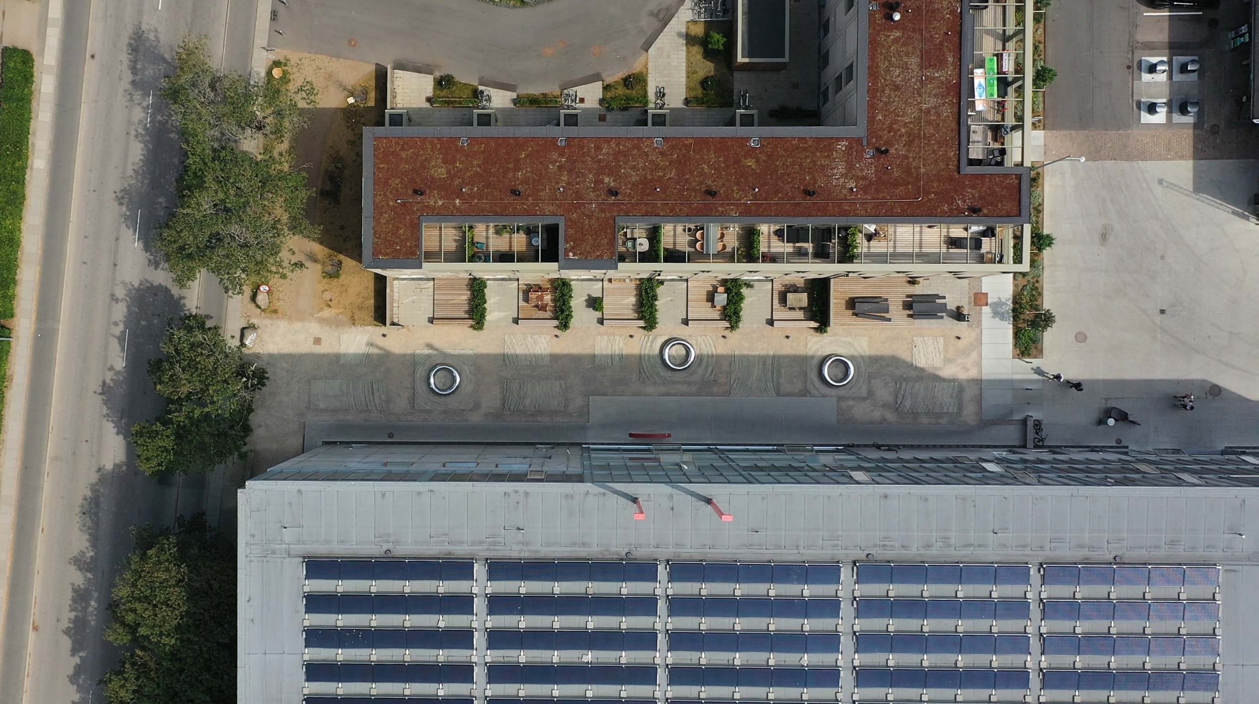 Aerial view af en urban boligkompleks med tagterrasse, planter, rendegraver og cykler, omgivet af bygninger, veje og træer.