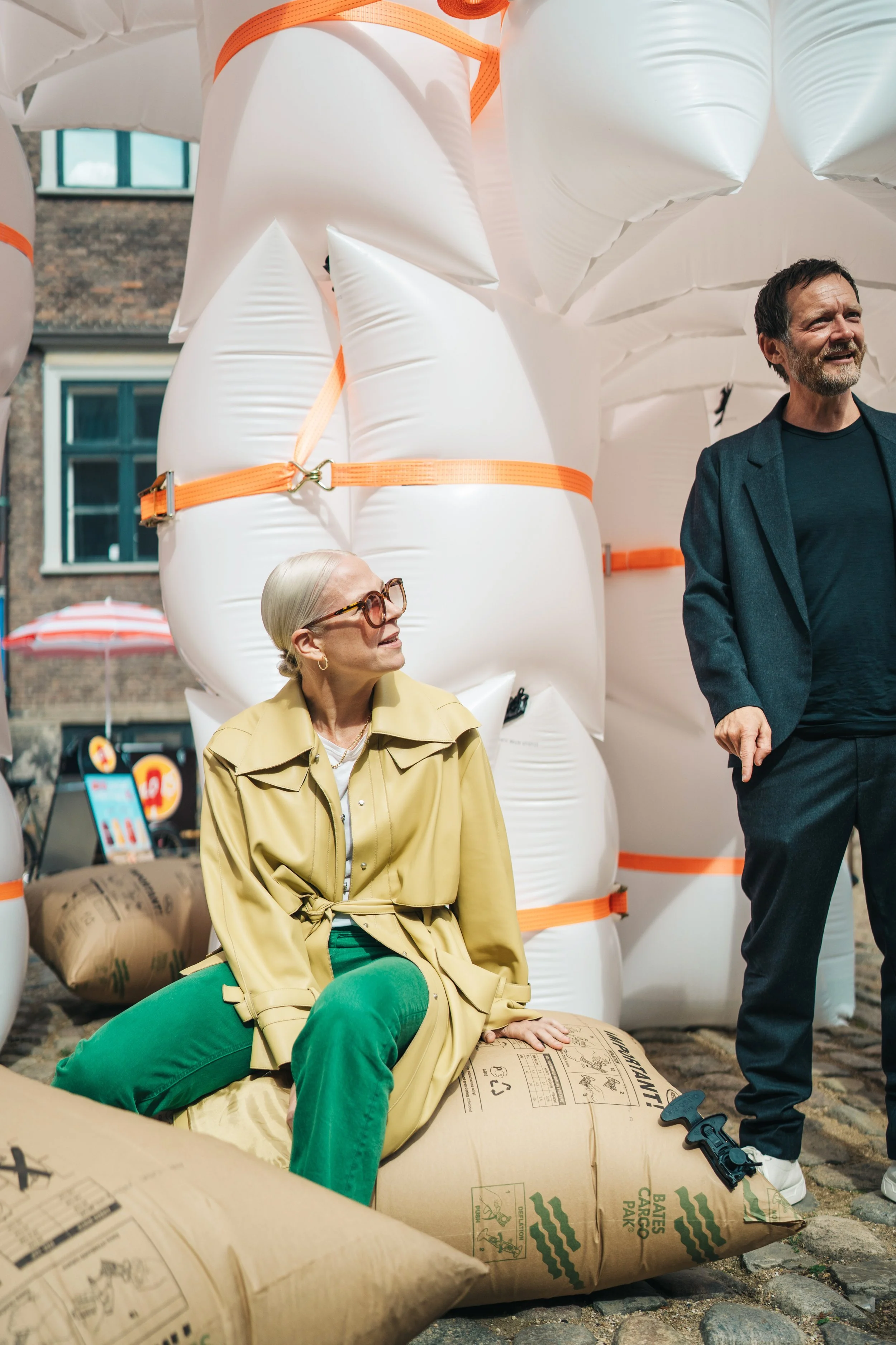 En ældre kvinde med blondt hår, briller og en beige trenchcoat sidder på en pose i en bygade, mens en mand står ved siden af. Bag dem er store oppustelige genstande bundet med orange bånd.