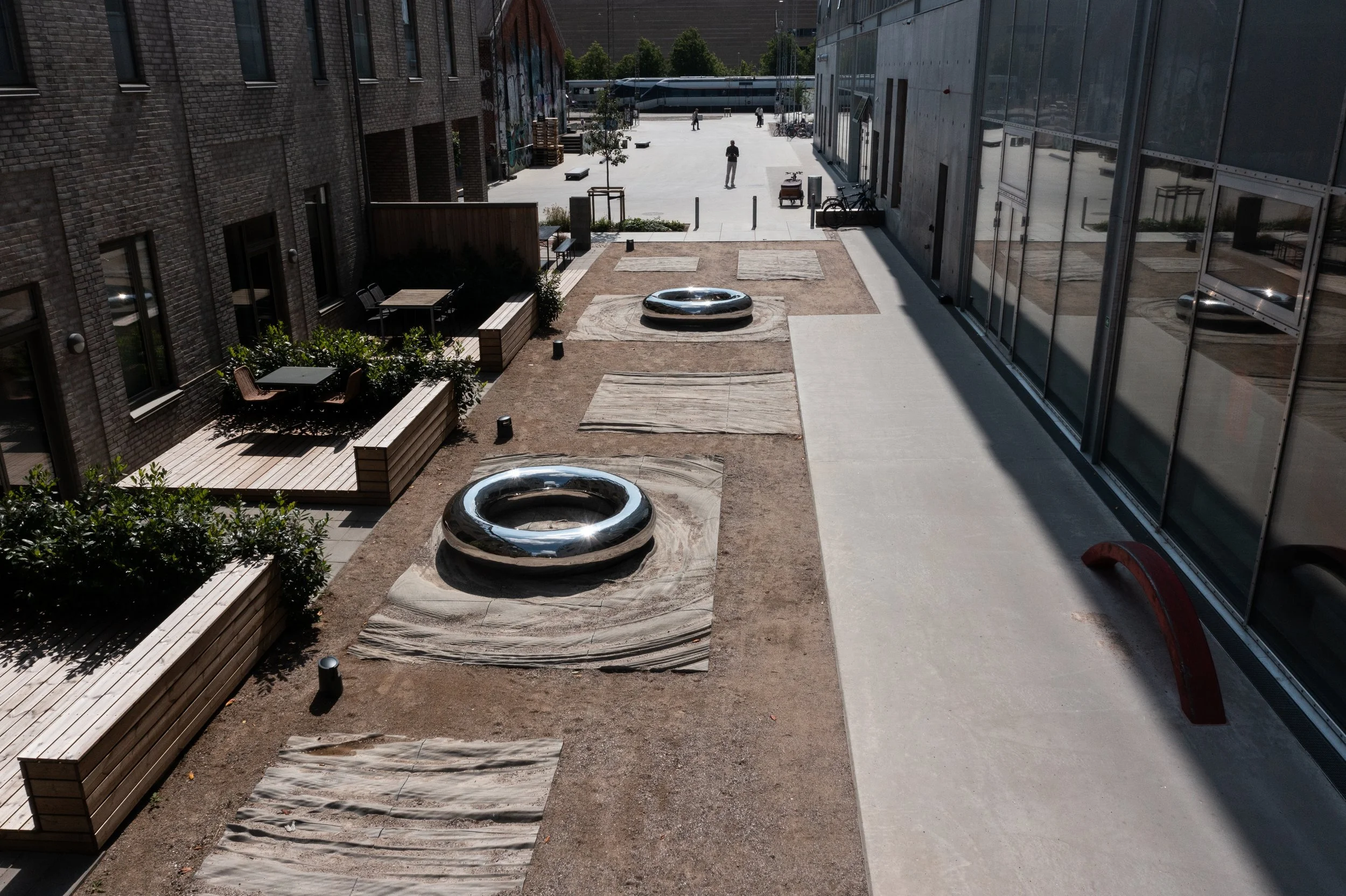 Fotografi af en moderne byudendørs plads med benkebænke, grønne planter og to dekorative metalring-formede vandsparekar, omgivet af bygninger og et åbent område i baggrunden.