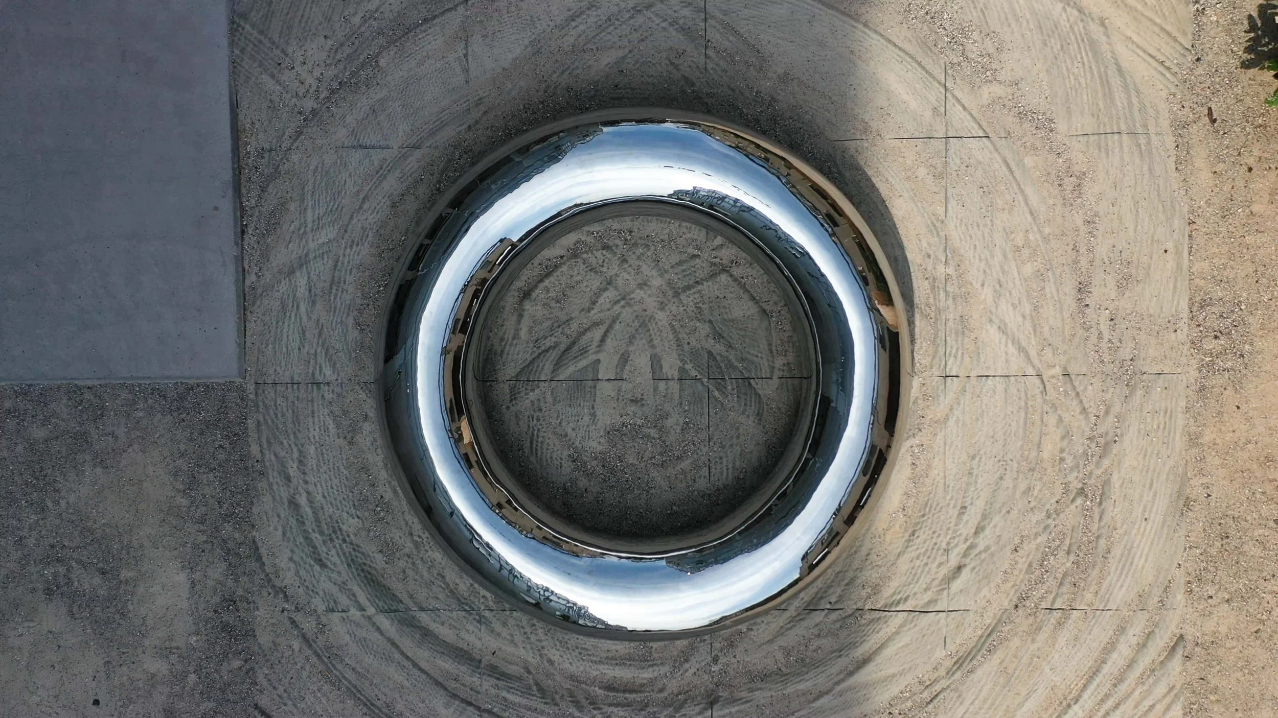 Refleksion i et rundmetalstykke på jorden, omgivet af grus og betonflise.