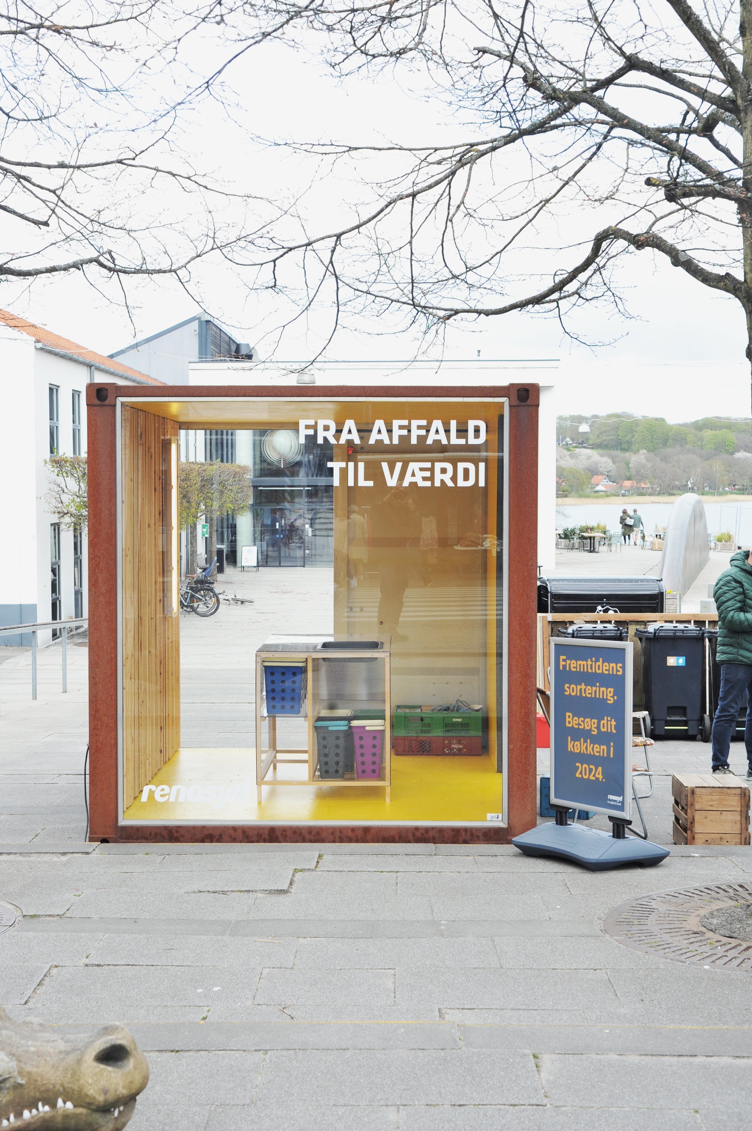 En genbrugsboks med teksten "Fra affald til værdi" på et offentligt sted. Der er en reklameskilt ved siden af med teksten "Fremtidens sortering. Besøg dit køkken i 2024."
