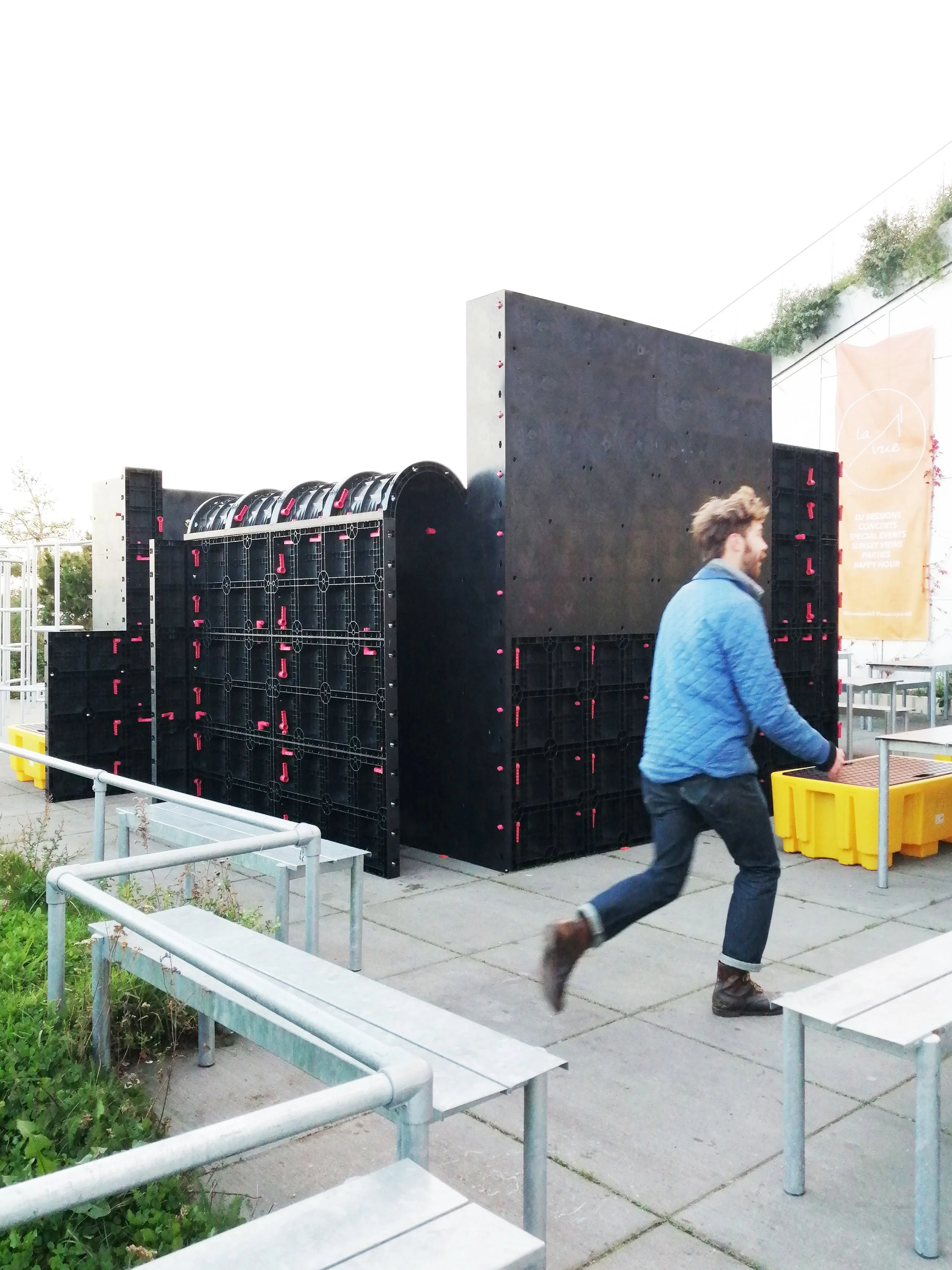 En mand løber foran en stor, sort struktur på en udendørs plads med grønne planter og metalrækker.