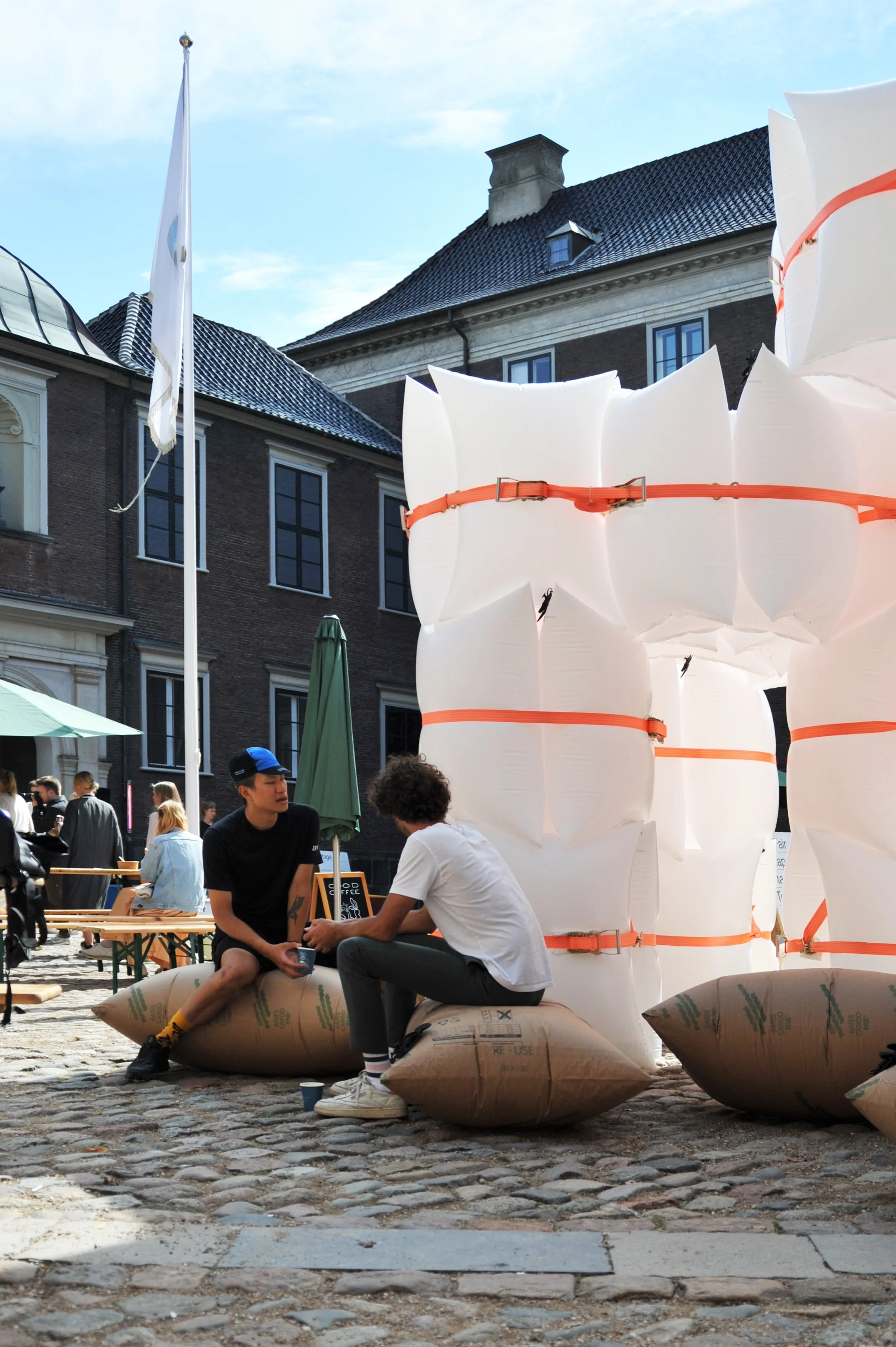 To unge mænd sidder på store puffedeil i en stor byrummiljø, omgivet af store oppustelige installationer og tager en samtale. Flere mennesker sidder på cafe-bænke i baggrunden.