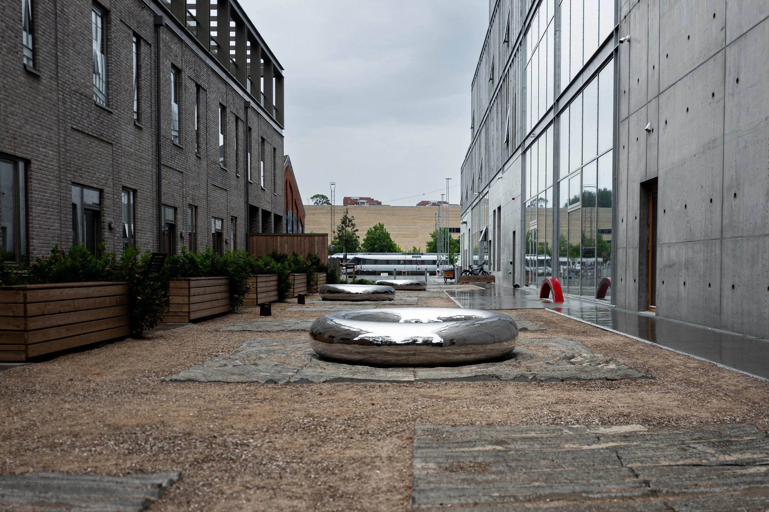 En moderne bygningsfacade med store refleksionsvinduer og et udendørs område med sten, træfliser og skinnende metalobjecter, under en overskyet himmel.
