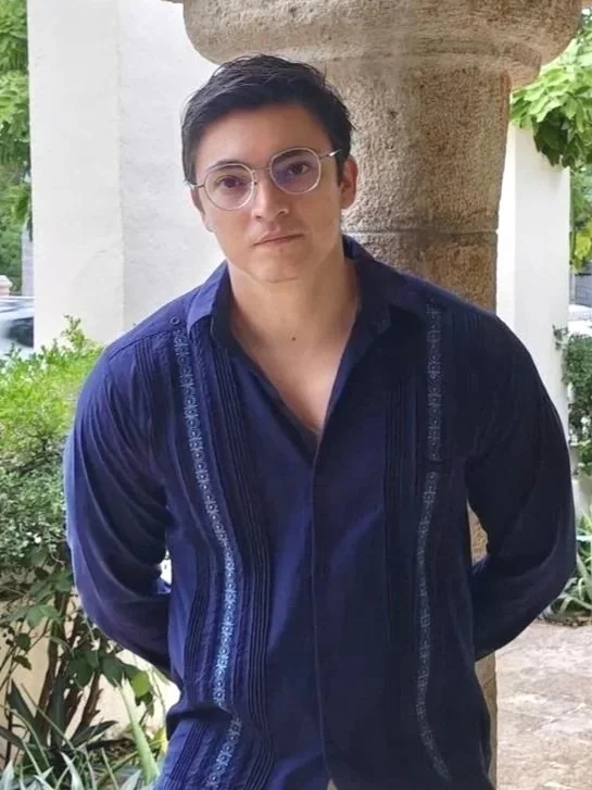 Joven con gafas, cabello oscuro y ropa azul, posa frente a una columna de piedra y vegetación.