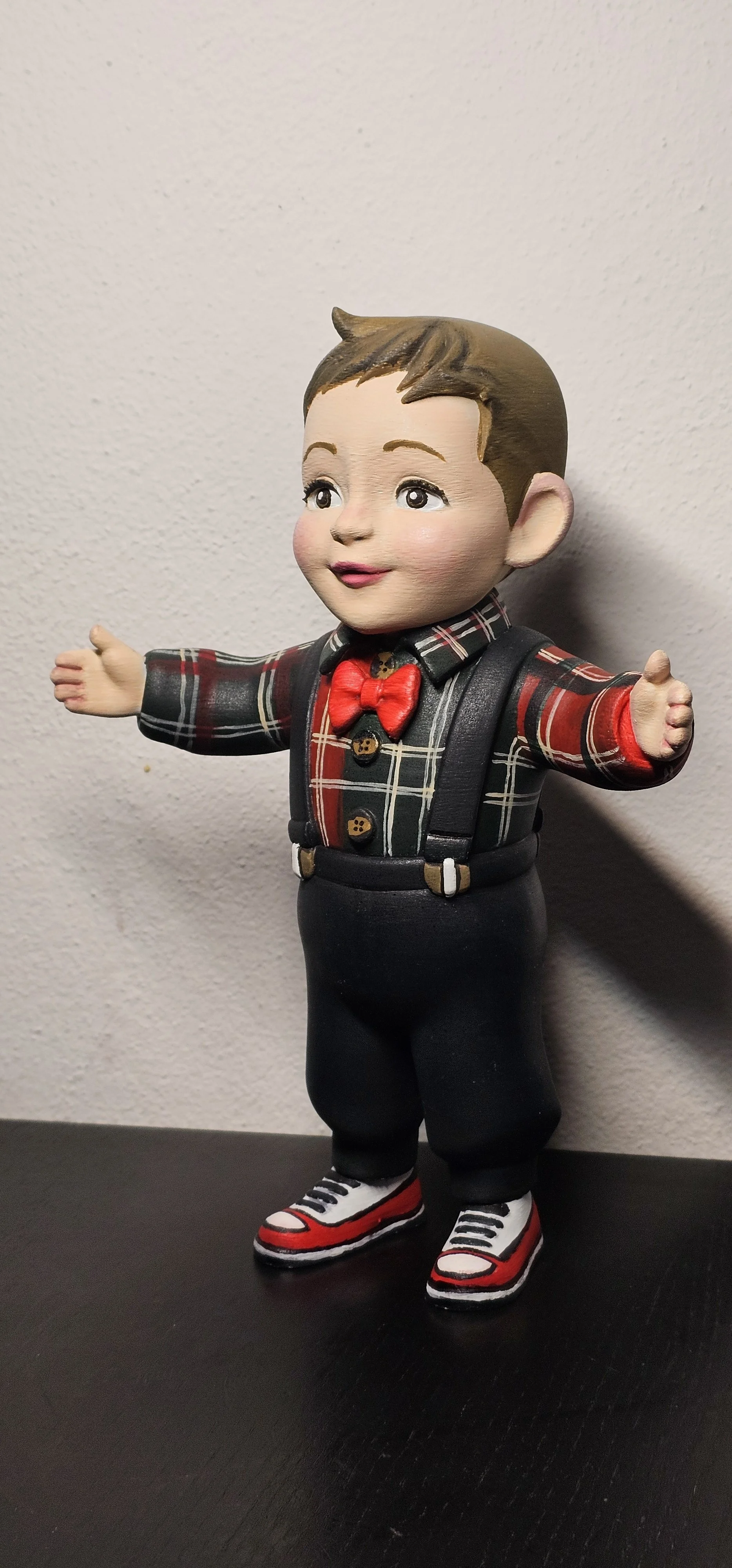 Statua di un bambino con braccia aperte, indossa una camicia a scacchi rossa, nera e beige, un papillon rosso, pantaloni neri, scarpe da ginnastica rosse, nere e bianche e uno zaino nero.