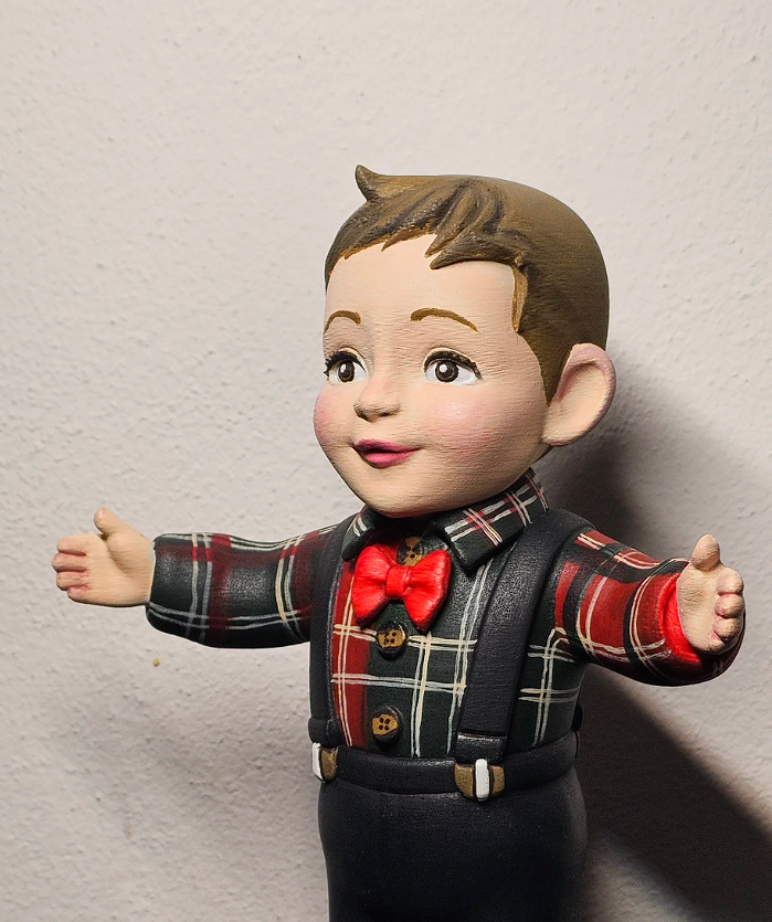 Statua di un bambino con le braccia aperte, vestito con una camicia a quadri rossa e nero, cravatta rossa, e bretelle nere, con una faccia sorridente e capelli castani.