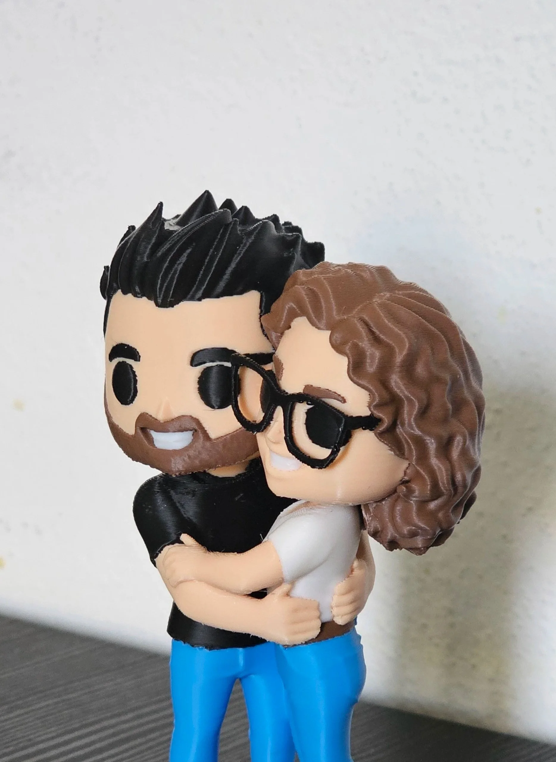 Statue di una coppia che si abbraccia, entrambi con occhiali neri, uomo con capelli e barba neri, donna con capelli ricci e castani. Entrambi sorridono. funkopop