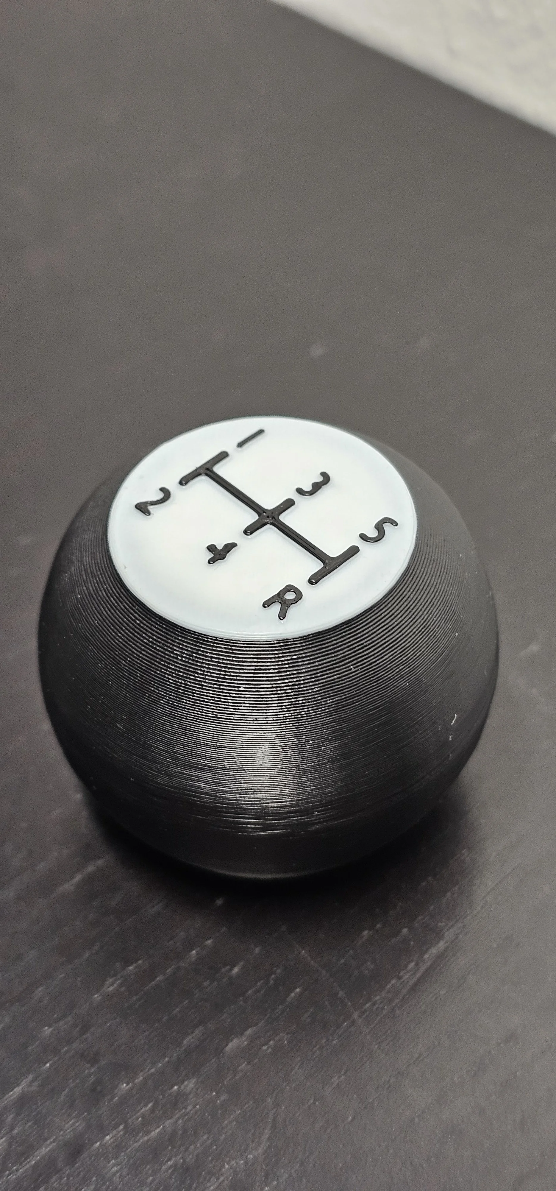 Un pomello del cambio di un'auto con la classica disposizione a H per le marce. La superficie è nera con una parte superiore bianca.