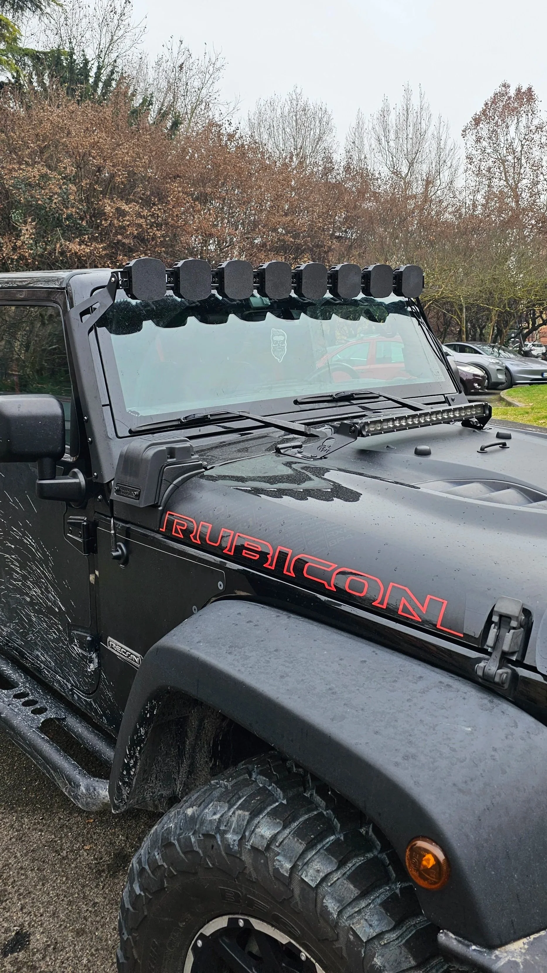 Un Jeep Rubicon nero dotato di luci su robusto tetto, con bersagli di fango sul fianco e un paio di alberi spogli sullo sfondo.