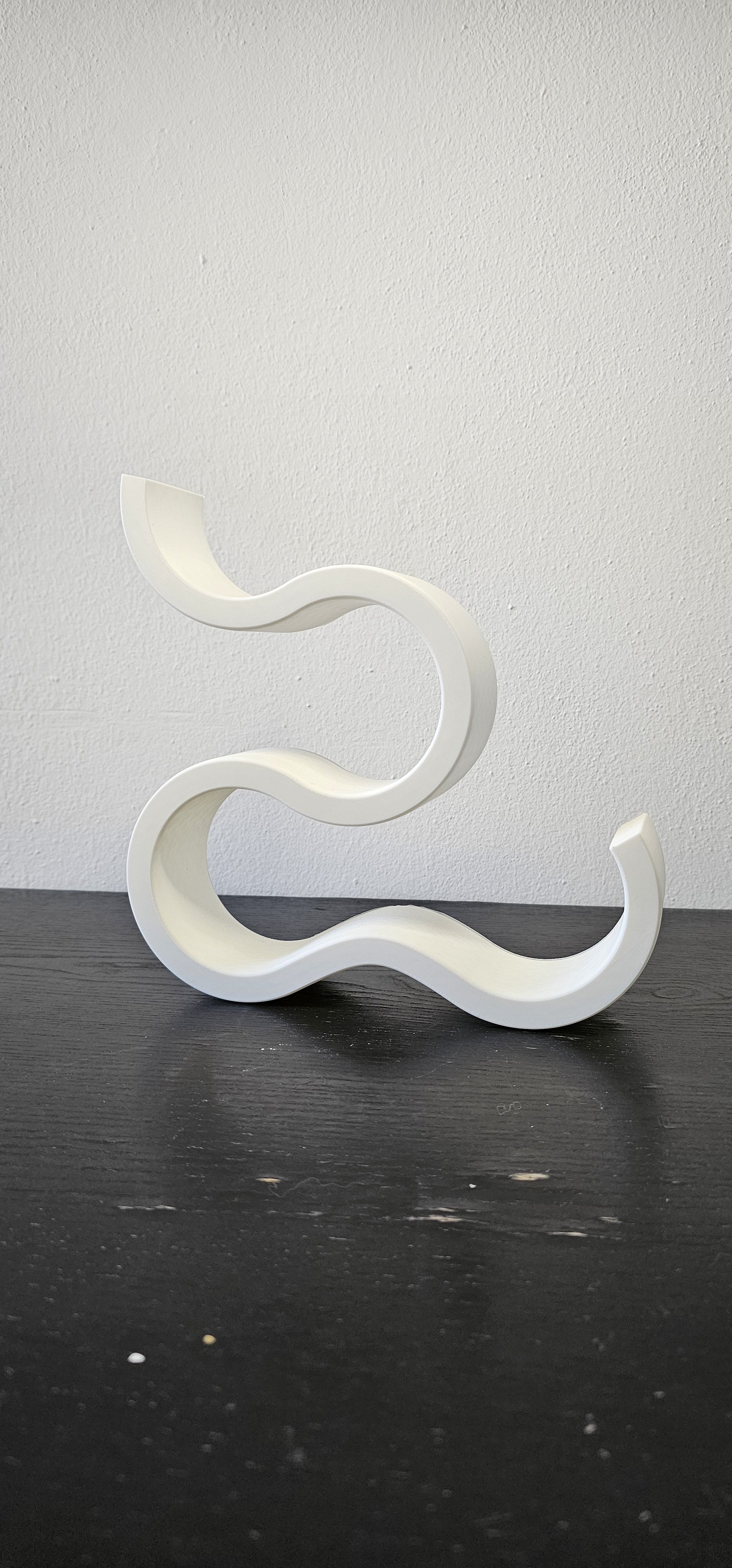 Scultura moderna bianca a forma di S curva porta vino, posizionata su un tavolo nero, con sfondo di parete bianca.