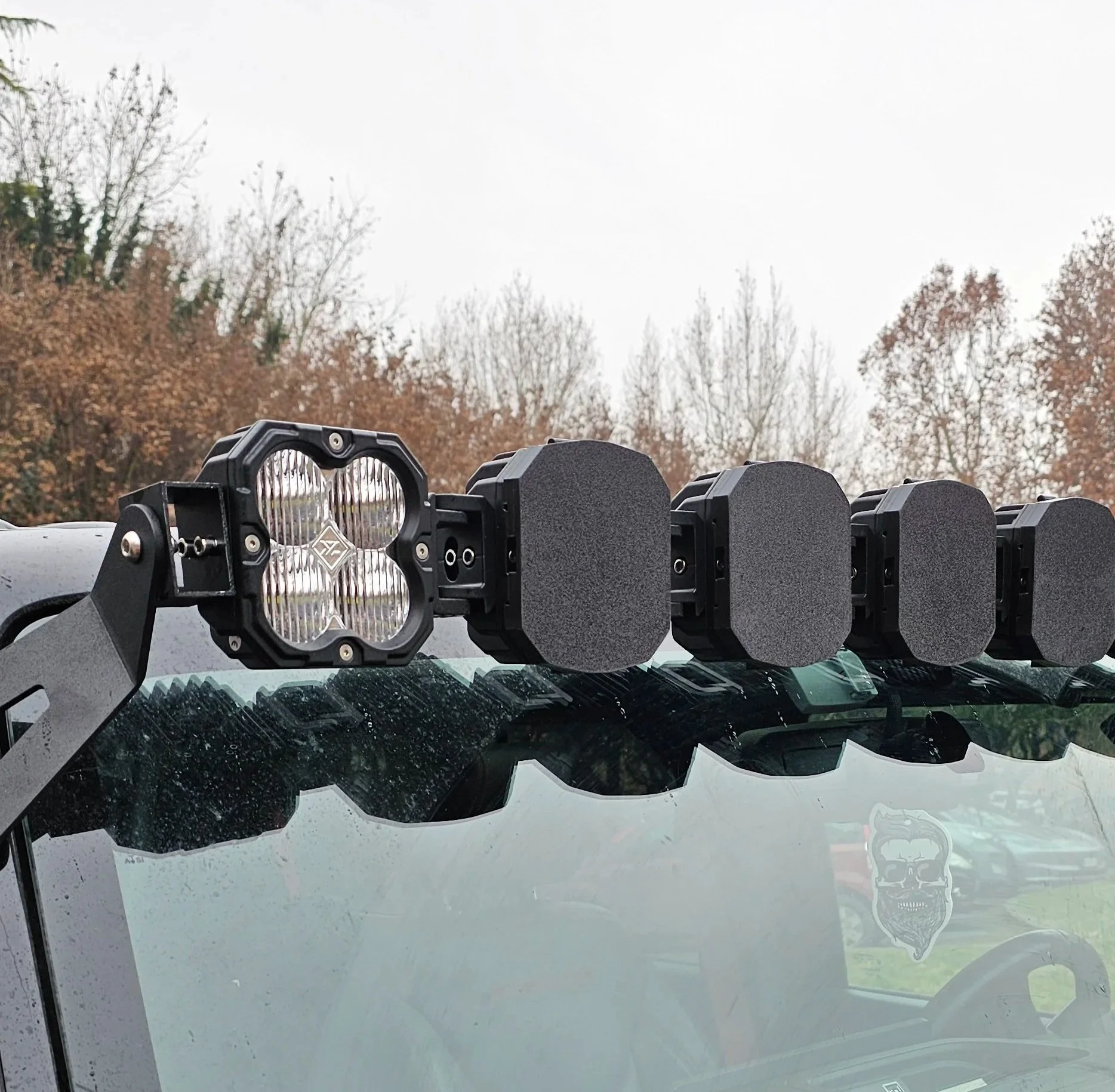 Faro LED da lavoro con quattro luci posizionato sopra il parabrezza di un veicolo, con un paesaggio di alberi spogli sullo sfondo.