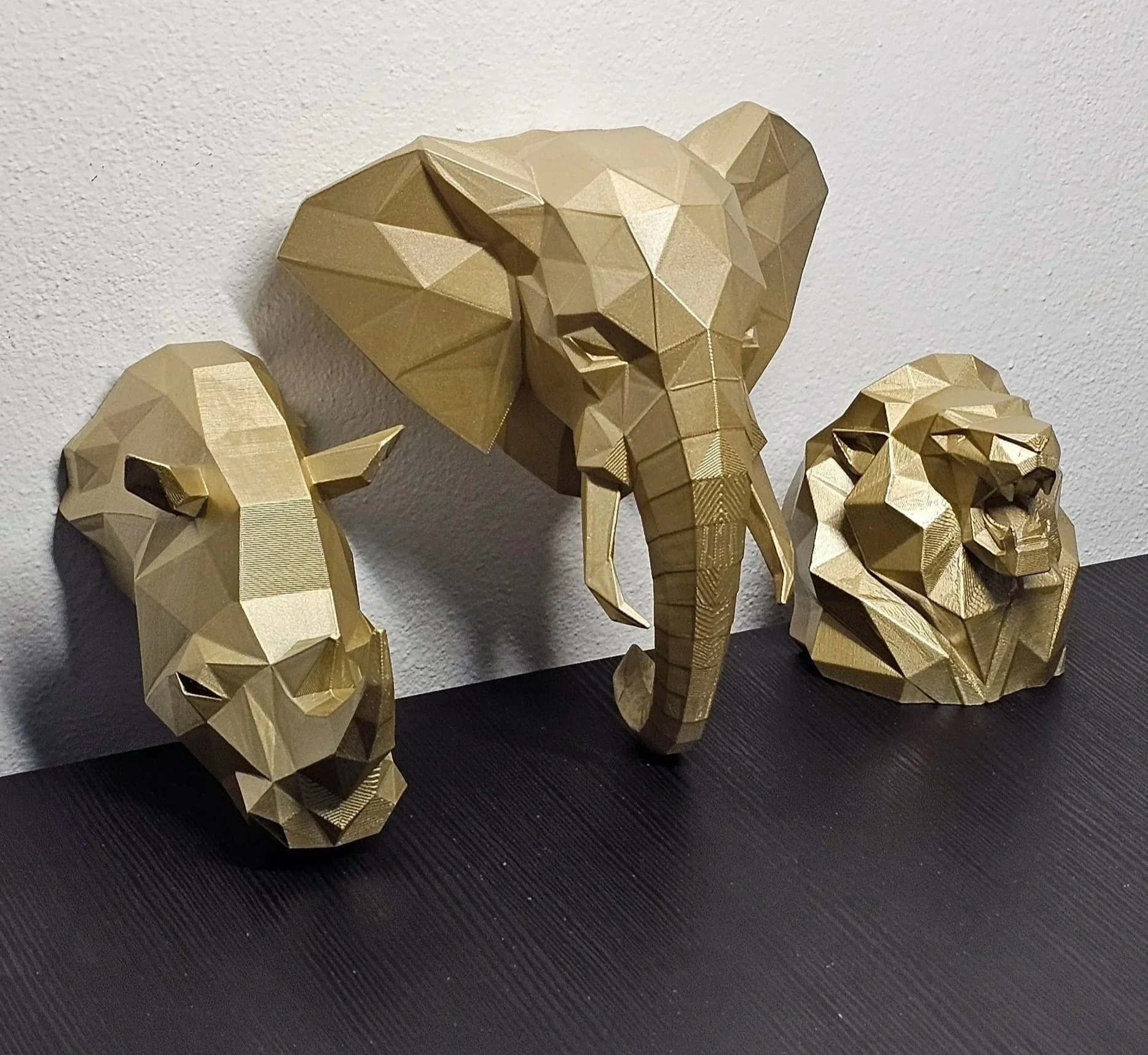 Statue di tre animali in stile geometrico: elefante al centro e due leoni ai lati, tutte di colore oro, posizionate su una superficie nera con uno sfondo di parete bianca.