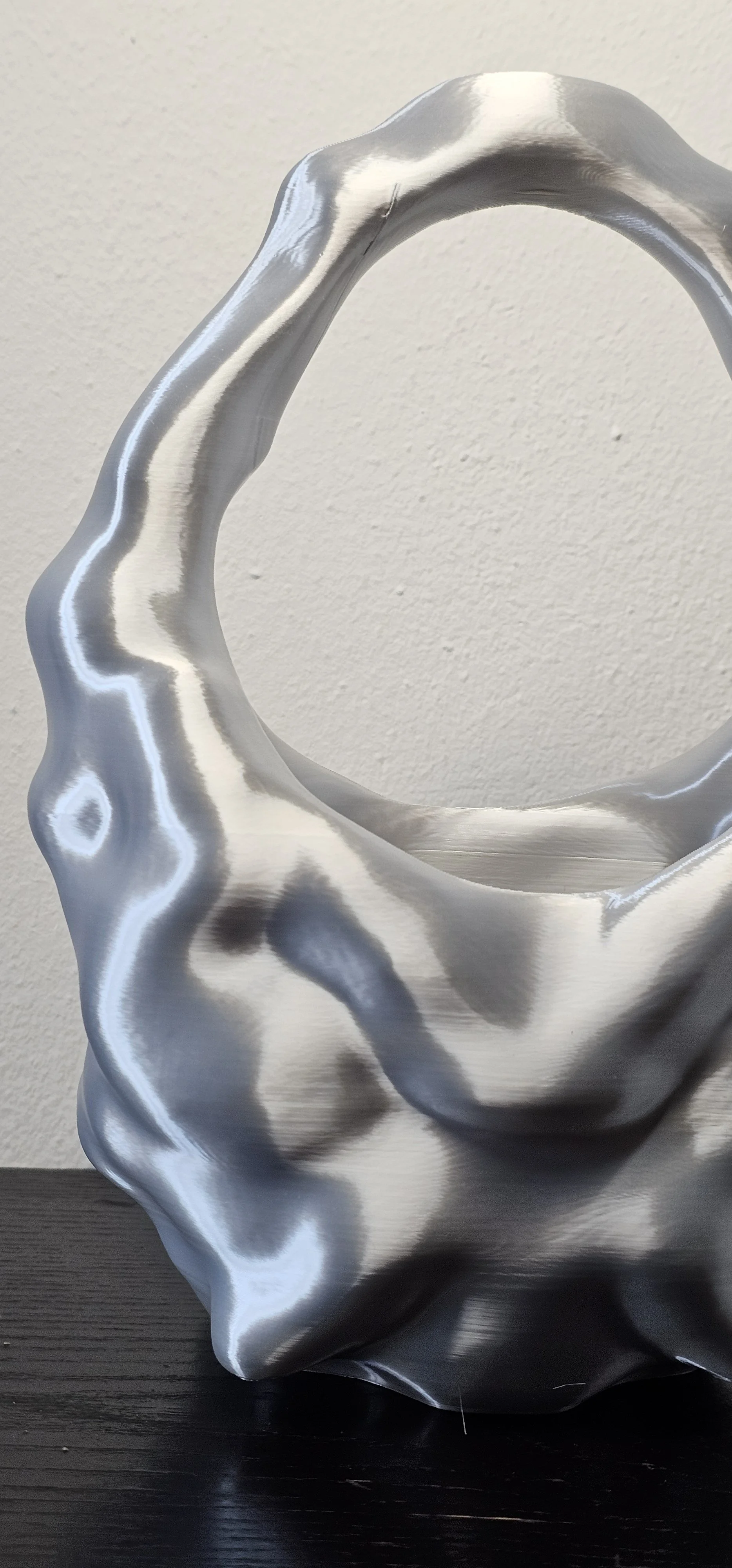 Scultura moderna in metallo a forma di borsa, superficie liscia e riflettente, posizionata su una superficie nera, sfondo bianco.