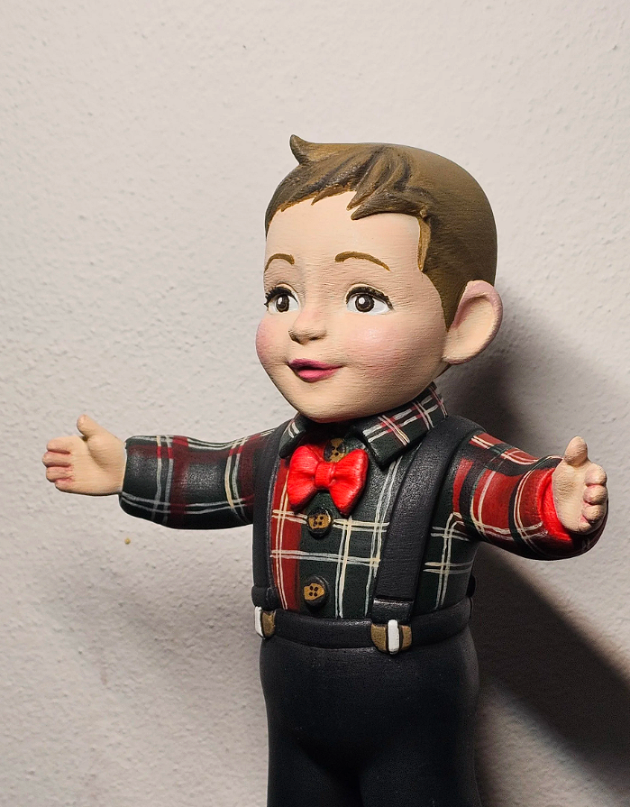 Statua di un bambino con capelli castani, volto sorridente, indossa una camicia a quadri rossa e nera con un fiocco rosso, pantaloni neri e bretelle nere, con le braccia aperte.