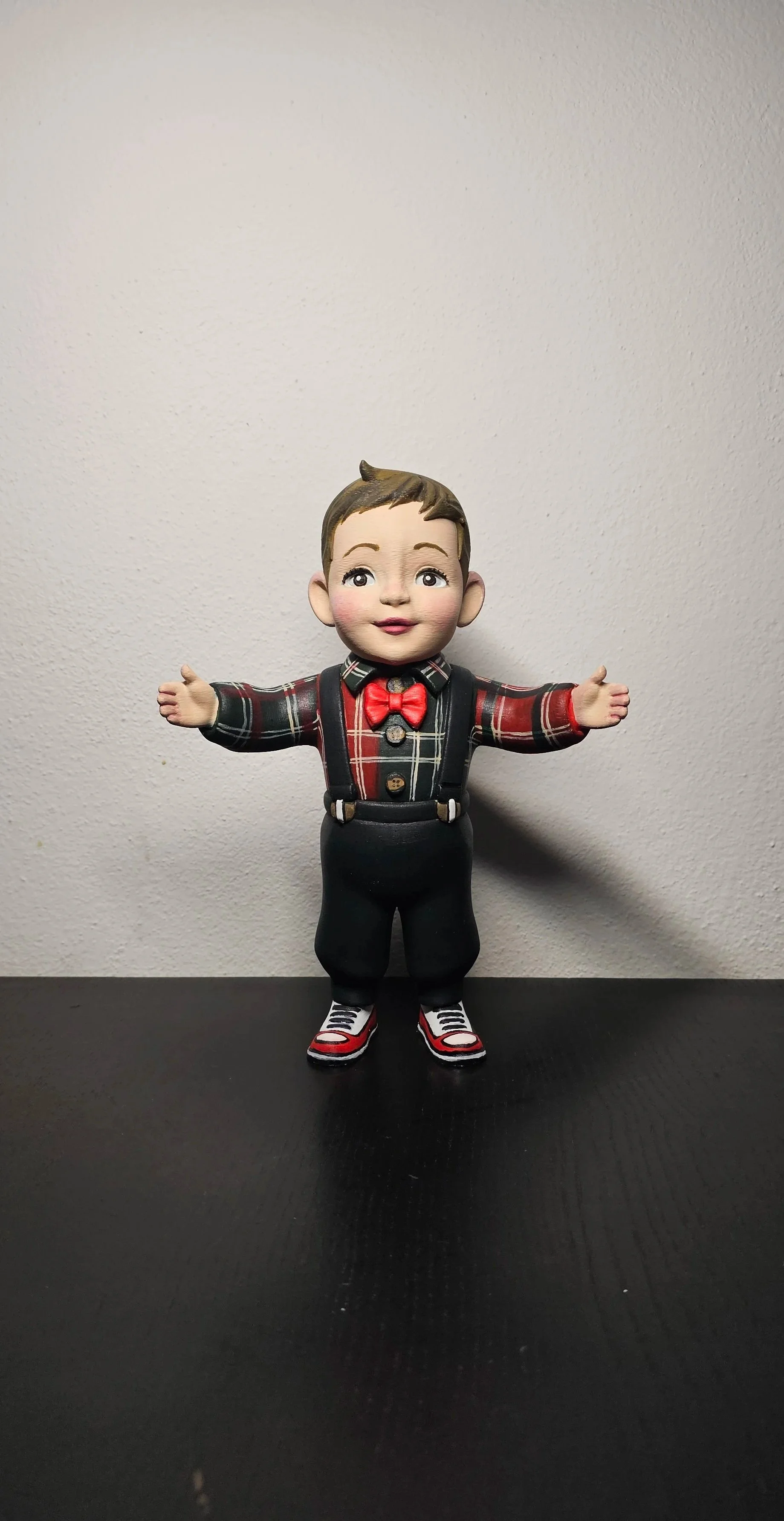 Statua di un bambino con braccia aperte, vestito con camicia a scacchi rossa e nera, con papillon rosso, pantaloni neri, scarpe da ginnastica rosse, nere e bianche, in piedi su una superficie nera contro una parete bianca.
