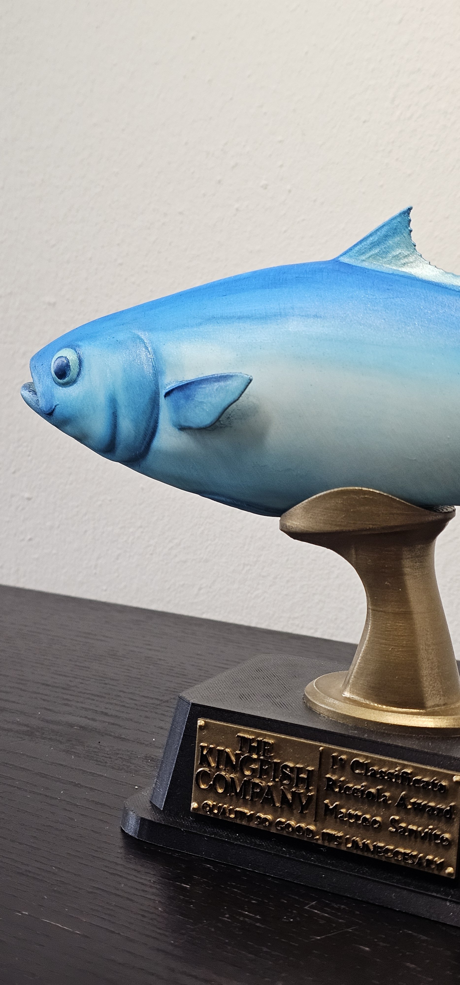 Trofeo a forma di pesce blu con pettorina bianca, montato su base di legno con placca di metallo con iscrizione appesa.