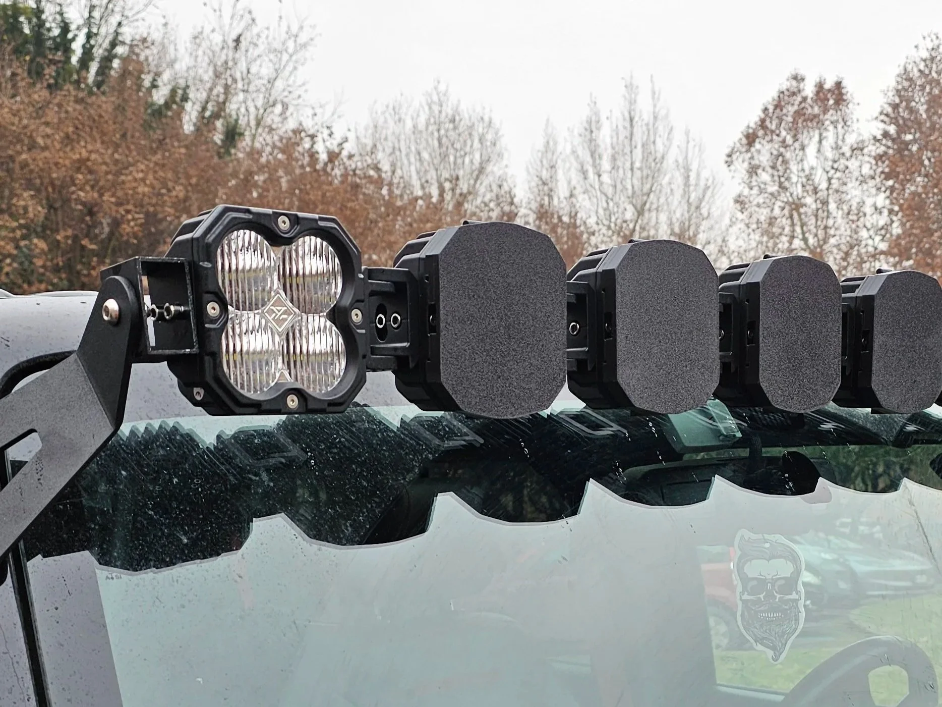 Fari da fuoristrada montati sulla parte superiore di un veicolo, con schermi di protezione neri e struttura robusta, con alberi spogli sullo sfondo.