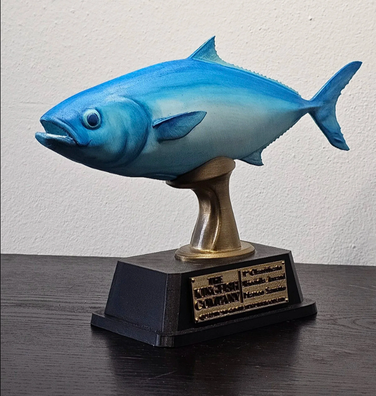 Una scultura di un pesce, di colore blu, montata su una base nera con una placca di metallo.