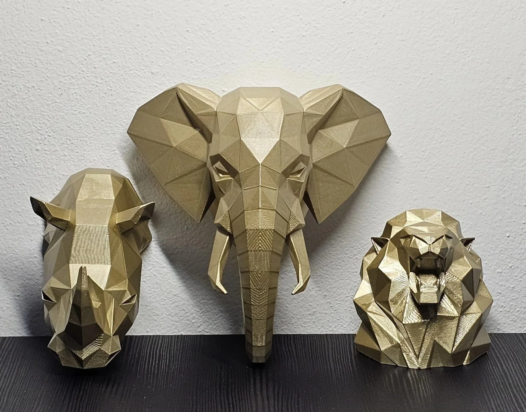 Tre sculture di animali fatte di materiali dorati con stile geometrico: un elefante centrale più grande e due leoni ai lati, uno sulla sinistra e uno sulla destra.