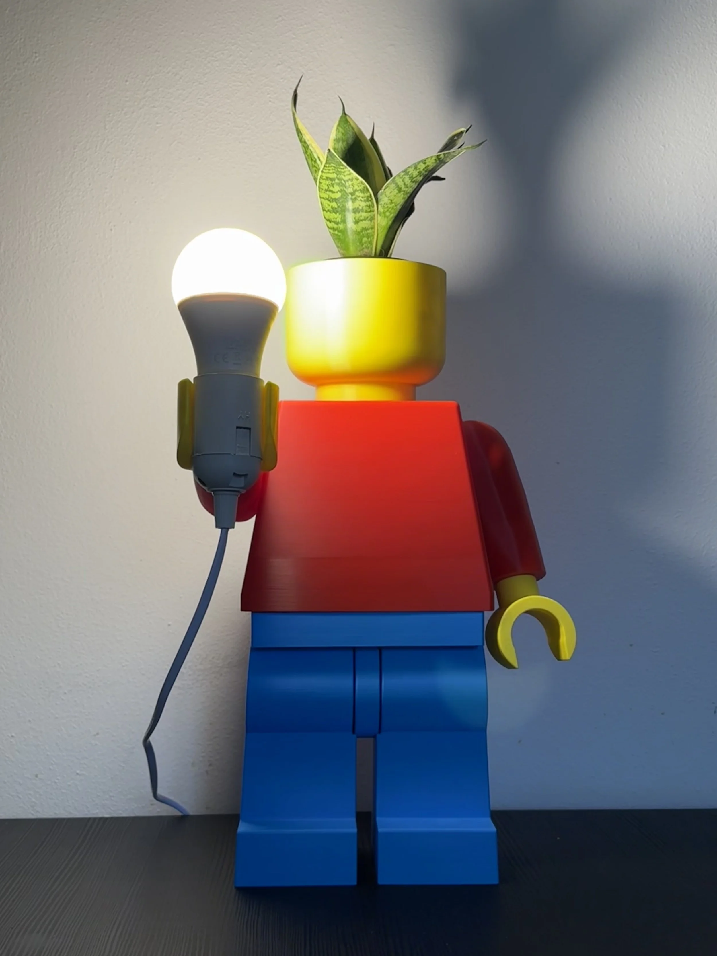 Statua di Lego rappresentante un personaggio con il corpo blu, maglietta rossa, braccia gialle, testa sostituita da una pianta in un vaso giallo, e che tiene in mano una lampadina accesa.