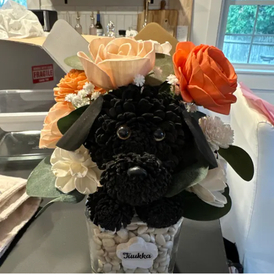 A black plush dog surrounded by peach, orange, and cream artificial flowers in a clear vase with white pebbles, labeled 'Tuulka.'