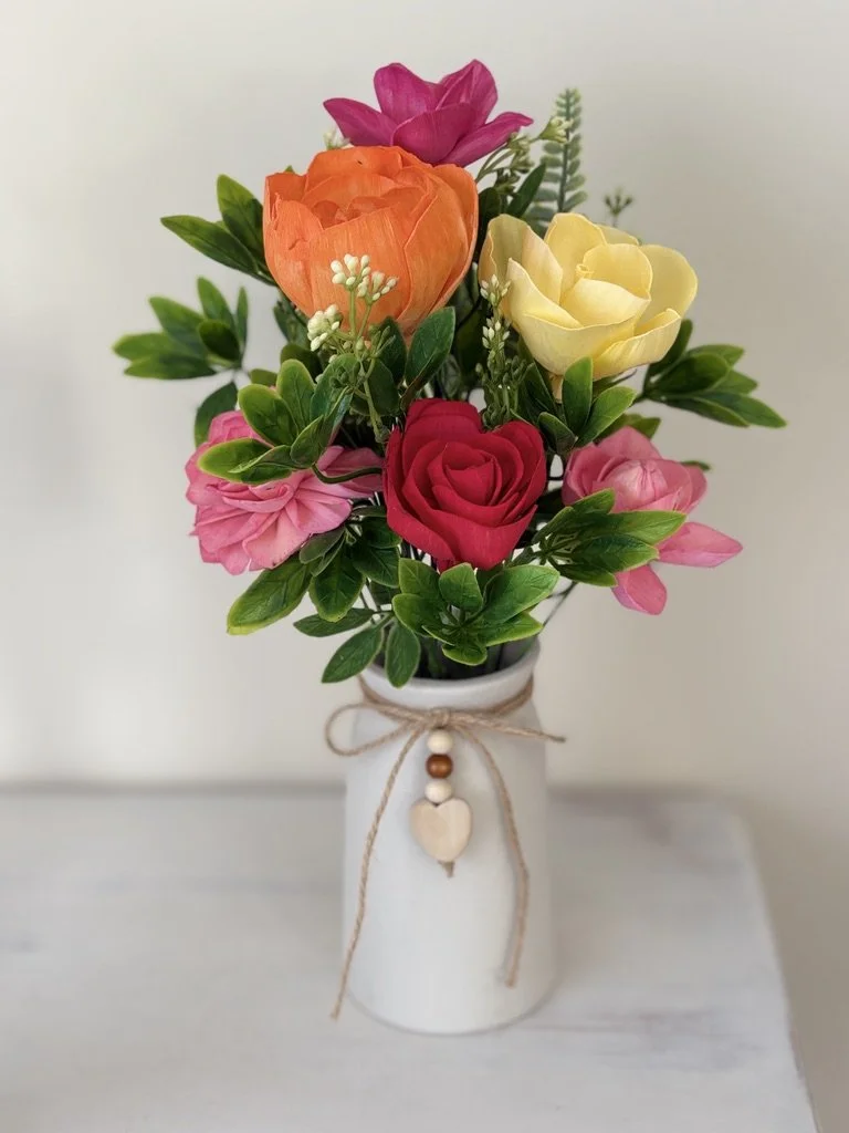 Colorful artificial flower bouquet with roses, ranunculus, and greenery in a white vase decorated with a twine bow and wood beads.
