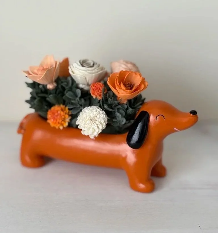A decorative planter in the shape of a dachshund dog with flowers inside its body, set on a neutral surface against a light background.