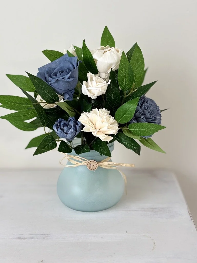 A light blue vase with a twine ribbon and wooden button, holding a bouquet of artificial flowers including blue and white roses and carnations, with green leaves, on a white surface.