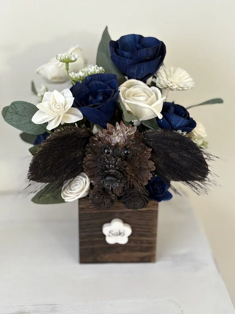 A floral arrangement featuring white and dark blue roses, white daisies, and greenery in a wooden box with a white label that says "Sabi." A small dog made from dark brown pinecones and wood accents is nestled among the flowers, with small googly eyes and a nose.