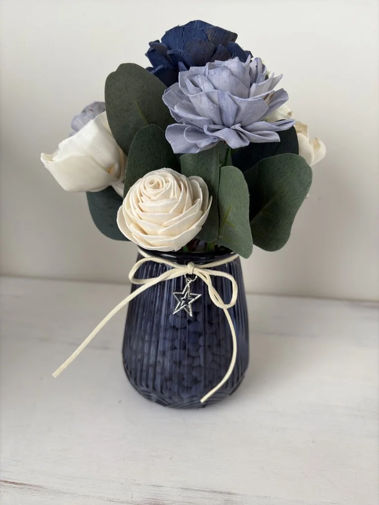 Decorative flower arrangement with white roses, blue and purple flowers, and green leaves in a dark blue vase with a cream ribbon and star charm.